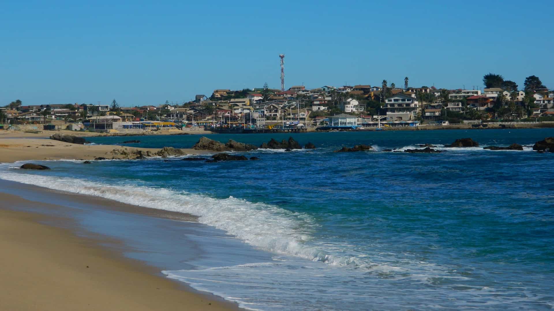 A detailed, horizontal photograph capturing a part of the beach and bay of the Coastal Town of El Quisco, Chile. The foreground features a wide stretch of golden sand leading to the bright turquoise Pacific Ocean, where small waves are breaking and washing over dark, rugged rocks. In the background, the colorful buildings, crowned by a tall communications tower, rise up a low hill overlooking the bay.