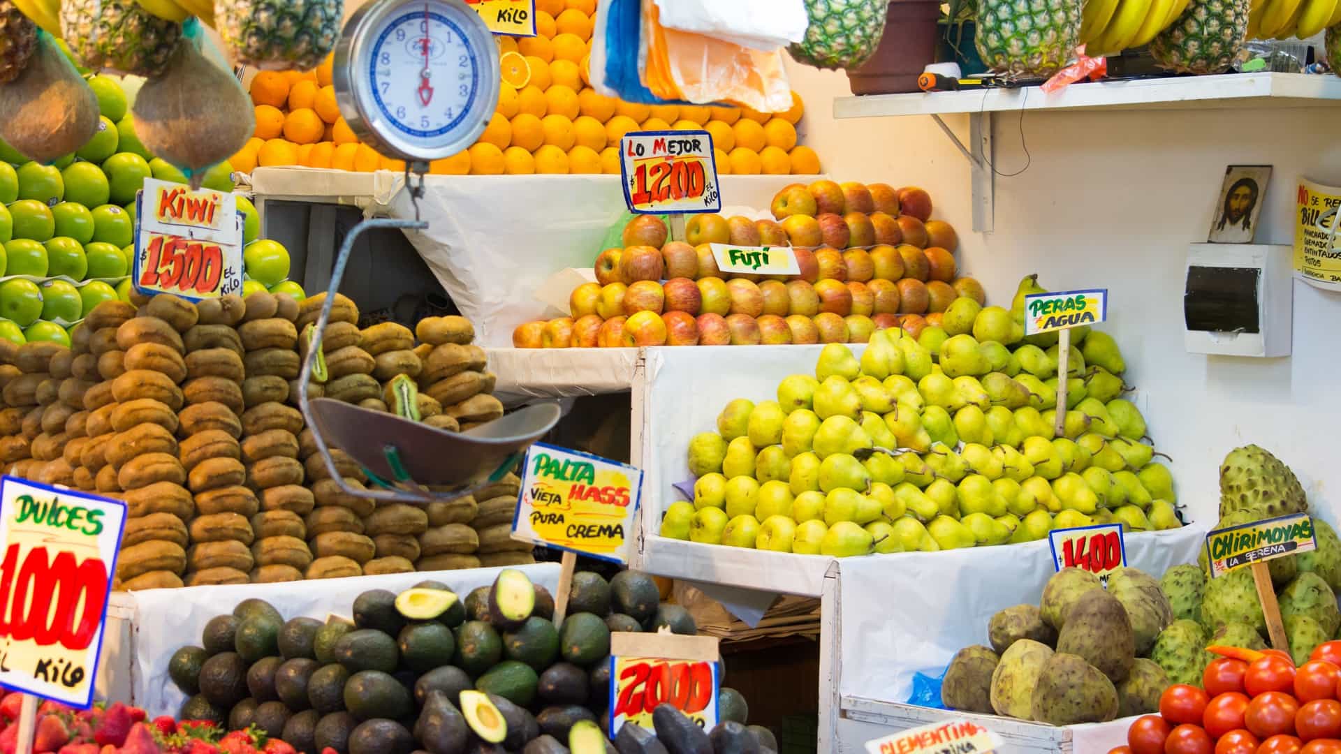 A close-up, colorful vertical shot of a vibrant, well-stocked fruit stall at the San Antonio Agricultural Market. The display is piled high with fresh produce, including neat stacks of green apples, yellow pears, kiwi fruit, avocados (palta), oranges, strawberries, cherimoyas (custard apples), and tomatoes.