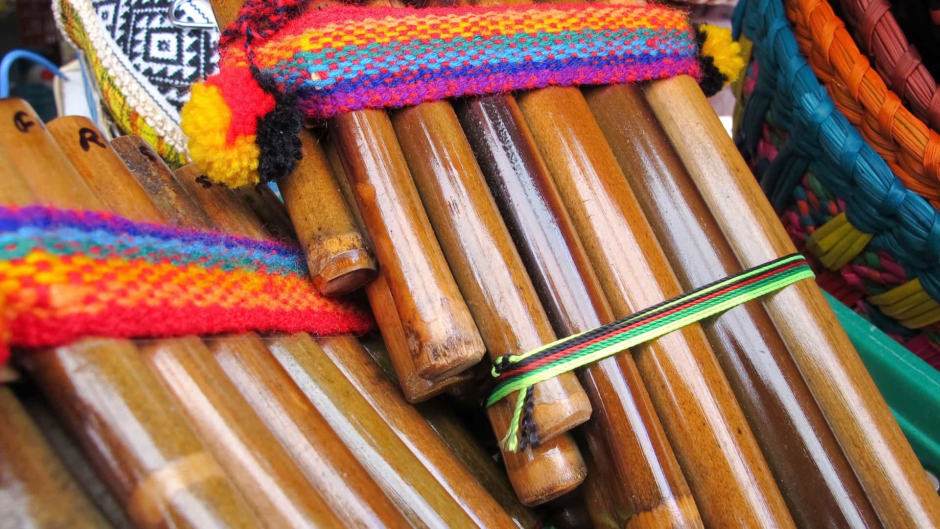 A close-up, warm-toned photograph showcasing a collection of South American pan flutes (sikus or zamponas) bundled together, displayed as a popular souvenir in the San Antonio/Santiago, Chile region. The wooden bamboo pipes are tied with colorful, hand-woven Andean textiles featuring bright geometric patterns. In the background, other woven crafts and baskets with traditional designs are visible at what appears to be a local market stall.