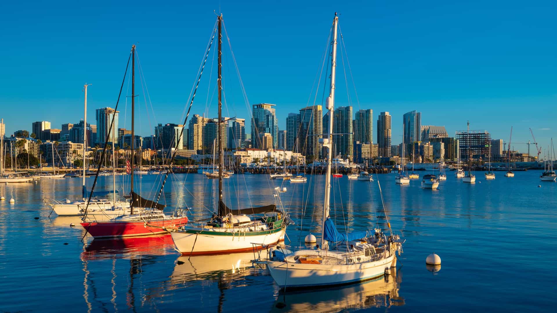 A dramatic sunrise or sunset photograph, likely taken from a boat harbor tour in San Diego, California, capturing three sailboats closely anchored. The harbor water reflects the vibrant blue sky and the boats' hulls, including one with a red hull and one with a white hull. In the background, the dense skyline of downtown San Diego is visible, silhouetted against the bright light, showcasing numerous modern skyscrapers.