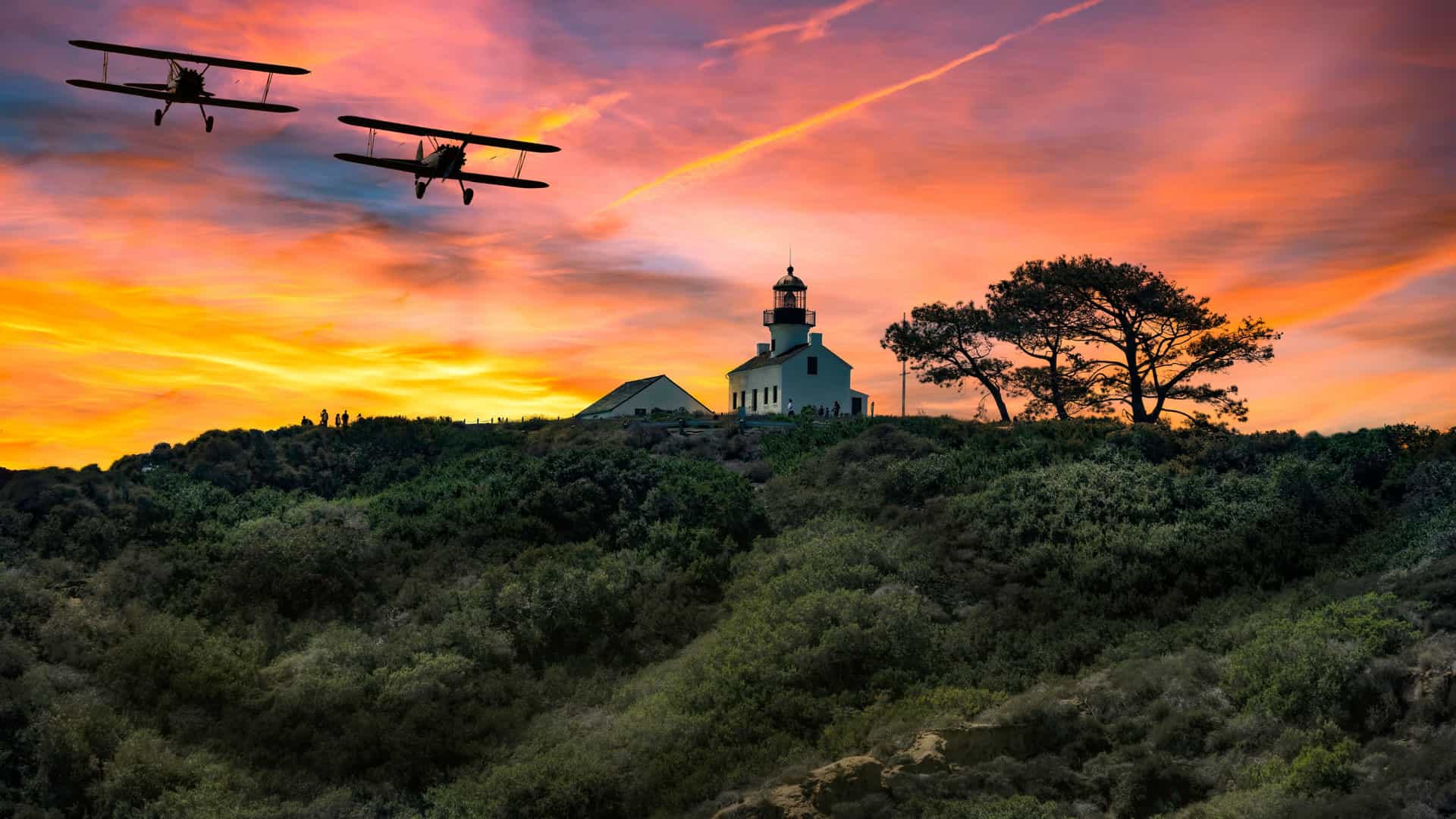 A dramatic sunset photograph, capturing the scene at the Cabrillo National Monument, showing the historic Old Point Loma Lighthouse situated atop a dark, brush-covered hill in San Diego. The sky is ablaze with intense hues of fiery orange, deep red, and gold. Two vintage biplanes are silhouetted in the upper left, flying across the intensely colored sky, leaving faint contrails. The lighthouse and a large, dark tree stand out sharply against the brilliant sunset.