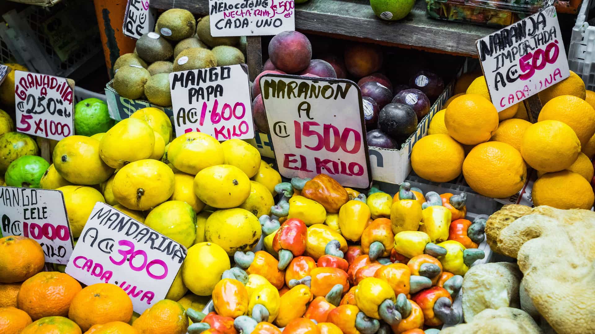 A close-up photograph of a colorful, overflowing market stall, likely located inside the famous Central Market (Mercado Central) in San Jose, Costa Rica. The display features a variety of tropical fruits, including yellow mangoes, oranges, and prominent clusters of bright red and yellow cashew apples (marañón). Handwritten price signs in Spanish are visible, using the Costa Rican Colón symbol (₡), indicating prices for imported oranges, mandarins, kiwi, and granadilla.