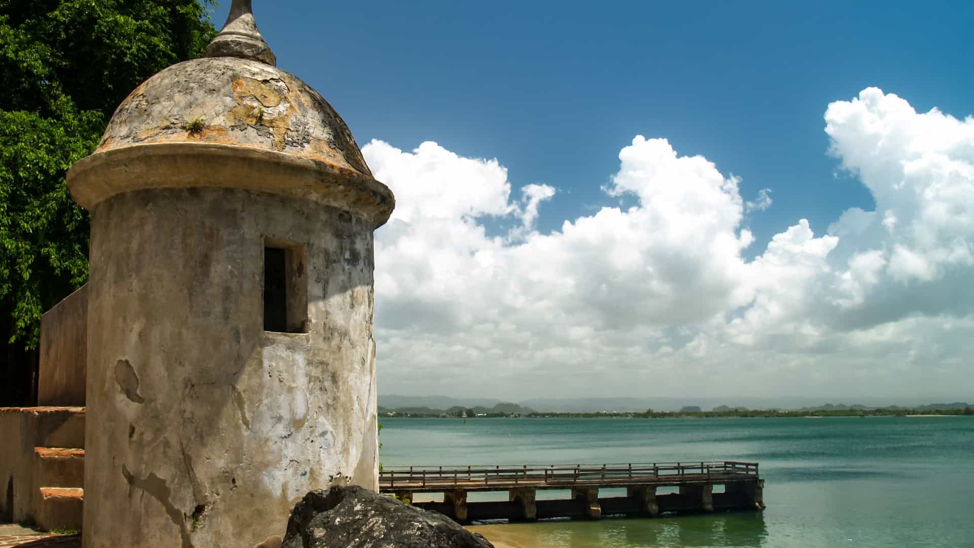  A close up view of a historic Spanish colonial garita (sentry box/watchtower) at Castillo San Felipe del Morro in Old San Juan, Puerto Rico. The stone sentry box stands prominently on the fortress's wall, overlooking the vast, deep blue Atlantic Ocean with white waves crashing on the rocky shore below. The sky is bright blue with a few scattered white clouds. The structure is an iconic symbol of the historic defenses.