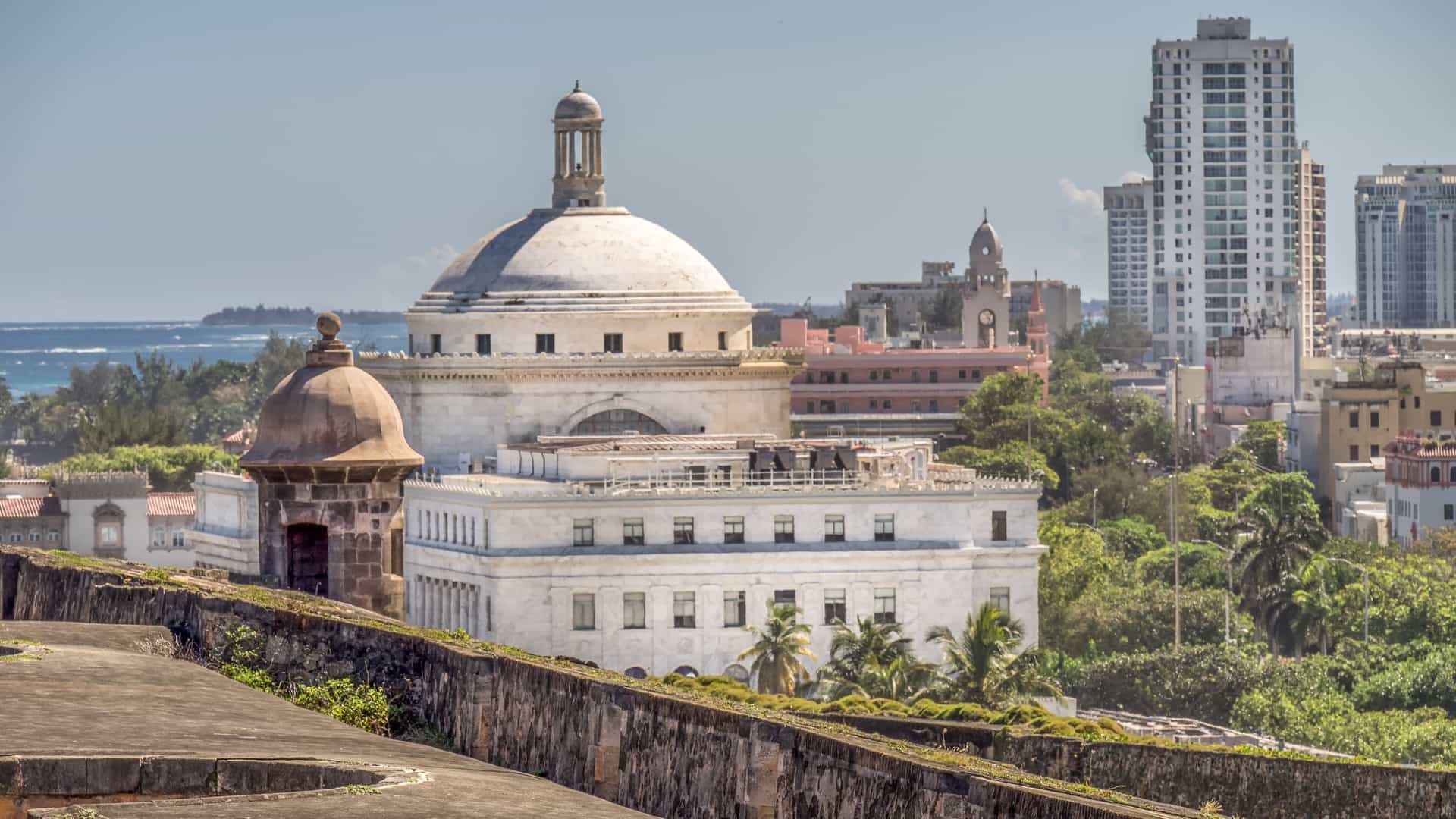  A view of the Castillo San Cristóbal, a massive Spanish colonial fortification in San Juan, Puerto Rico. The photo focuses on a section of the fort's stone outer walls and an integrated garita (sentry box) on the corner. The fortress is situated on a grassy hill with a view toward the ocean. The sky is bright blue with scattered white clouds, and the scene is sunlit.