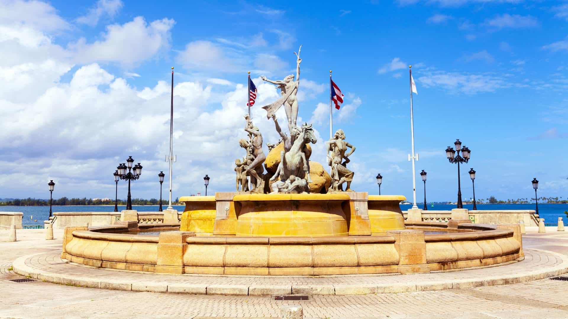  The Fuente Raíces (Roots Fountain), a large bronze sculpture located at the end of the Paseo de la Princesa promenade in Old San Juan, Puerto Rico. The statues depict figures symbolizing the Taíno, Spanish, and African heritage of the island, with water splashing around them and the San Juan Bay visible in the background.