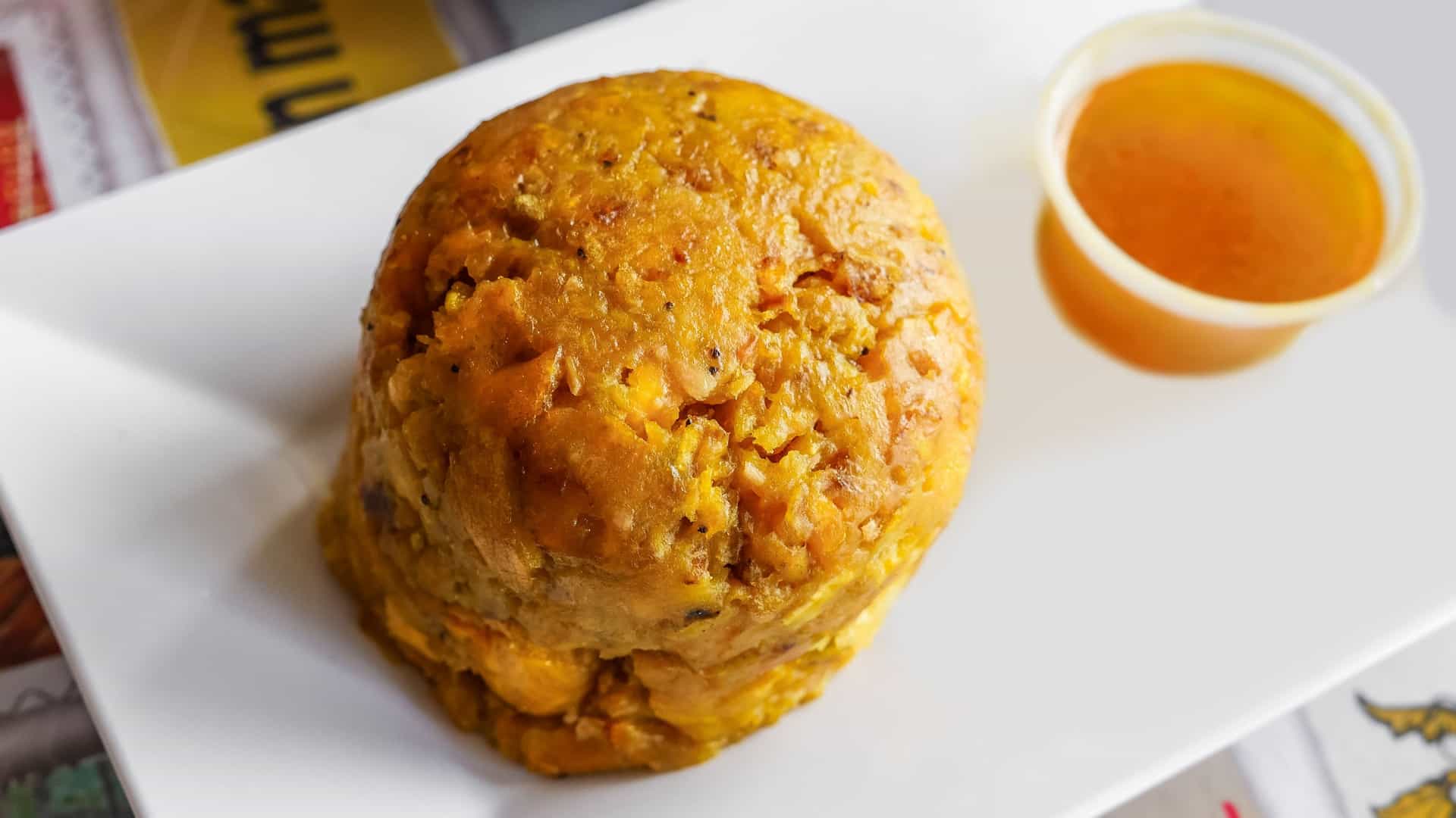  A close up of Mofongo, a traditional Puerto Rican dish. The mashed, dome shaped mound of fried green plantains, garlic, and pork cracklings (chicharrón) is served on a white plate, likely surrounded by a savory broth or sauce and garnished with fresh herbs.