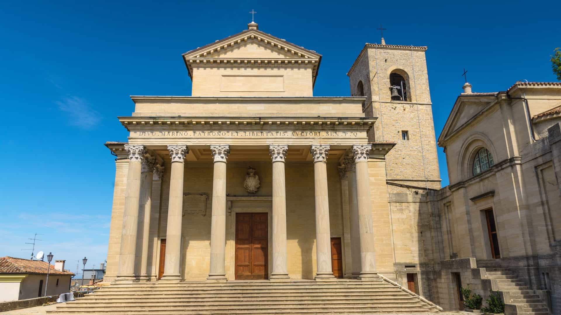  The exterior facade of the Basilica di San Marino, the principal church in the Republic of San Marino. The church is built in the Neoclassical style with a grand front porch (pronaos) featuring eight white Corinthian columns supporting a triangular pediment. A stone bell tower rises to the right of the main structure. The basilica is situated at the top of a wide staircase on Piazzale Domus Plebis under a bright sky.