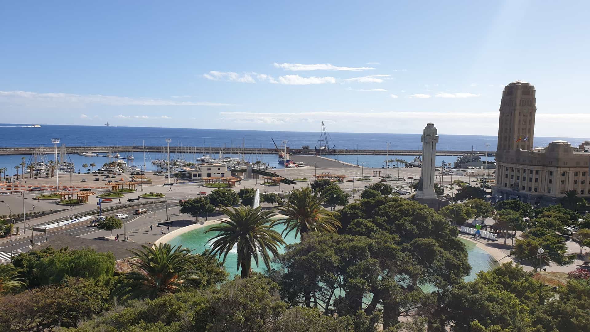  An elevated panoramic view over the city of Santa Cruz de Tenerife, Canary Islands, showing a dense collection of white and light colored buildings sloping down towards the large blue harbor and the Atlantic Ocean. The futuristic, wing shaped Auditorio de Tenerife is visible on the waterfront, with rugged mountains rising in the background.