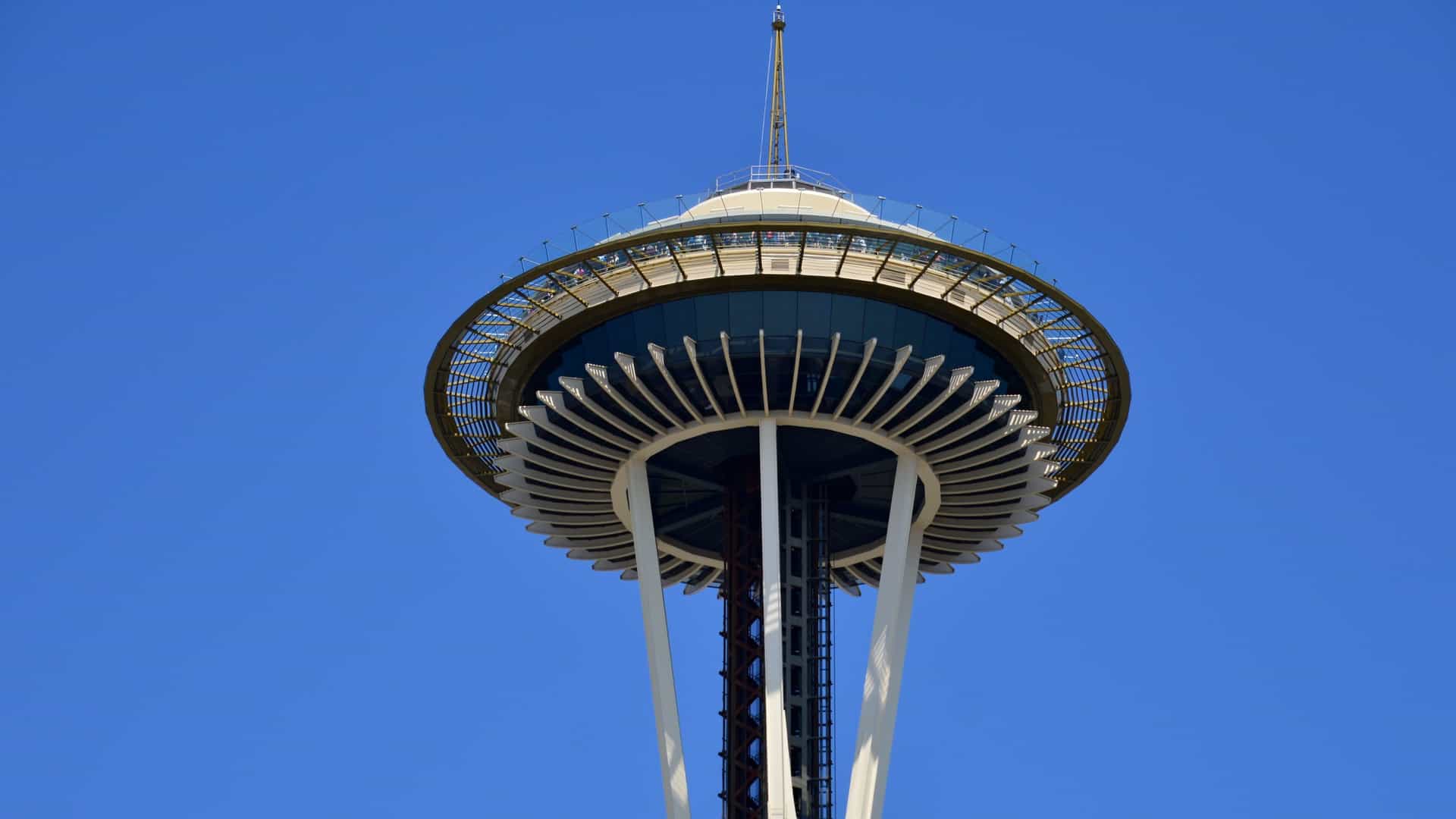 A clear, close-up view of the iconic Space Needle in Seattle, Washington, a slender tower with a saucer-shaped observation deck and rotating restaurant at the top. The structure stands prominently against a bright blue sky, showcasing its futuristic mid-century modern design.