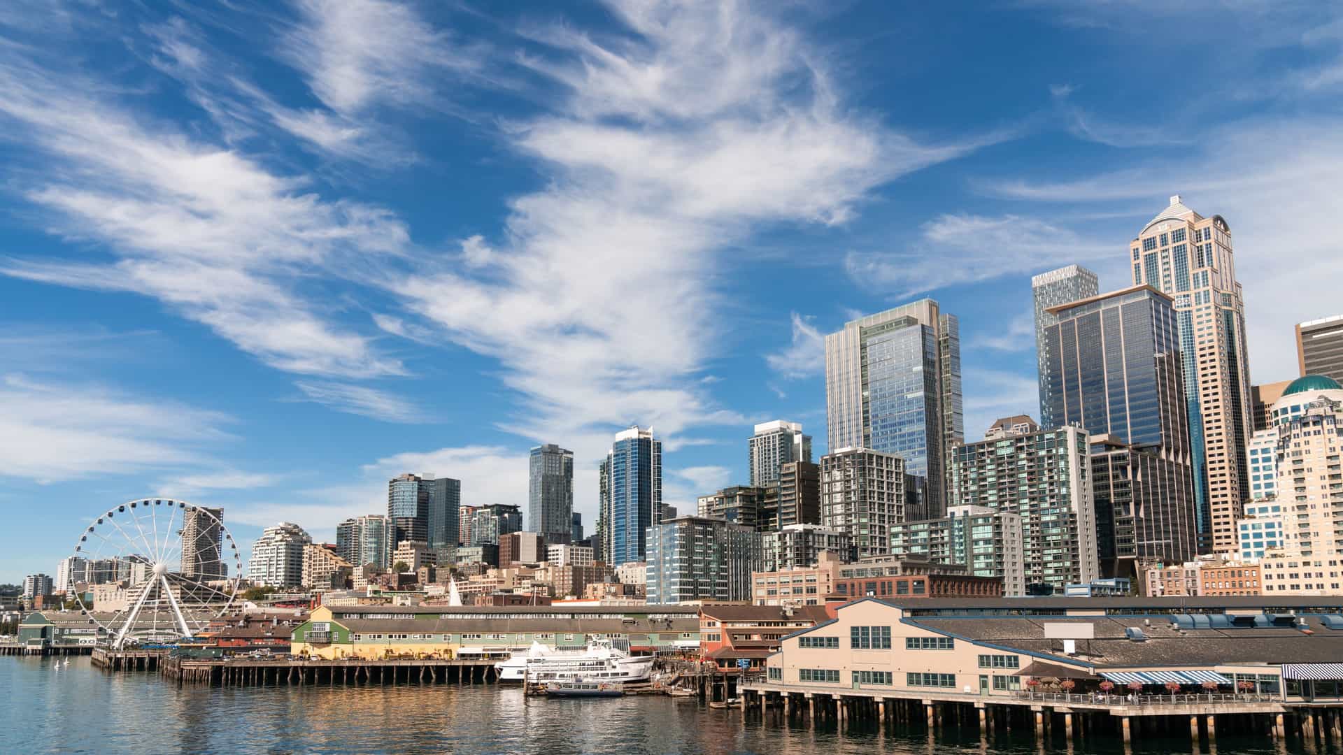 A scenic view of the Seattle waterfront and downtown skyline. In the foreground, the Seattle Great Wheel, a large, lit-up Ferris wheel, stands on a pier over Elliott Bay. Behind it, the dense cluster of downtown skyscrapers and office towers completes the iconic cityscape.