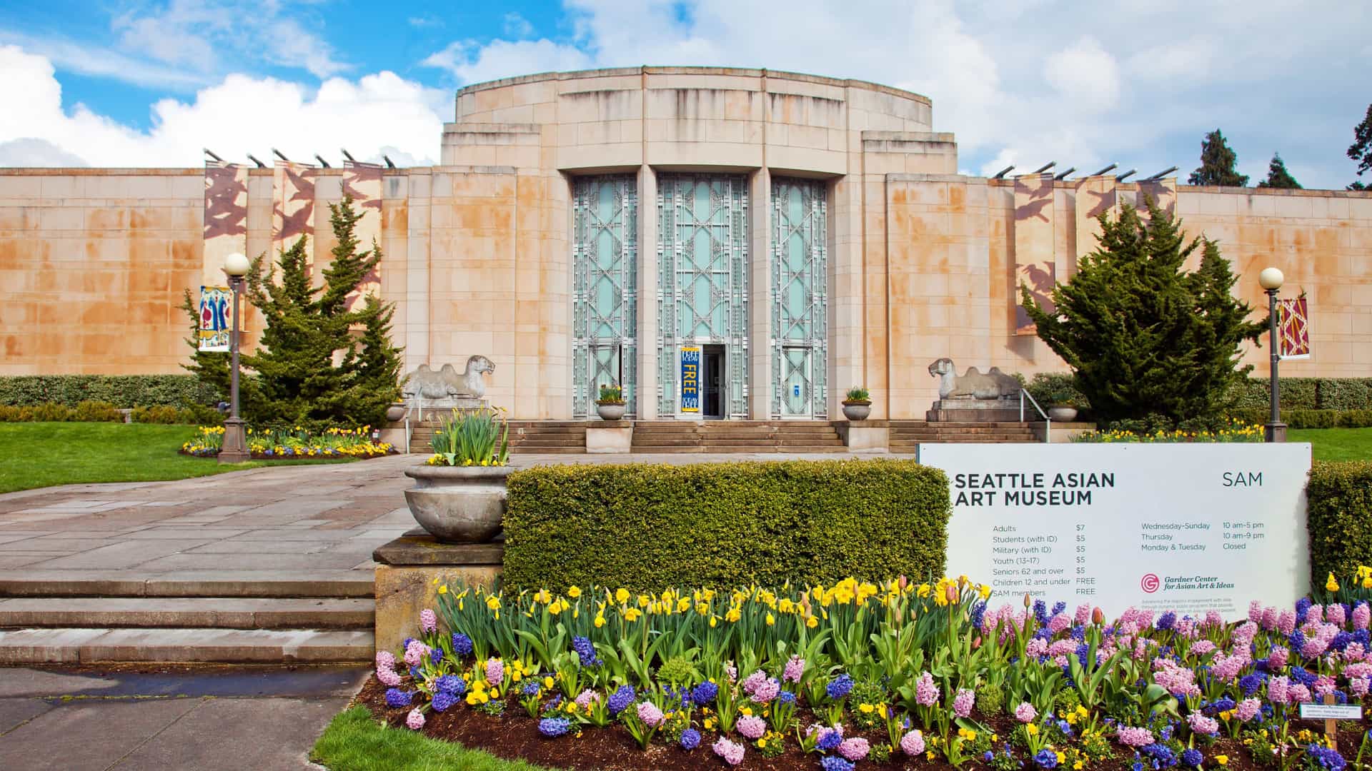 The Seattle Asian Art Museum, an elegant Art Deco building, is set within the lush grounds of Volunteer Park. The photograph captures the symmetrical facade of the historic structure, which features Wilkeson sandstone and Art Moderne architectural elements. The well-maintained foreground includes manicured lawns and garden beds leading up to the museum entrance.