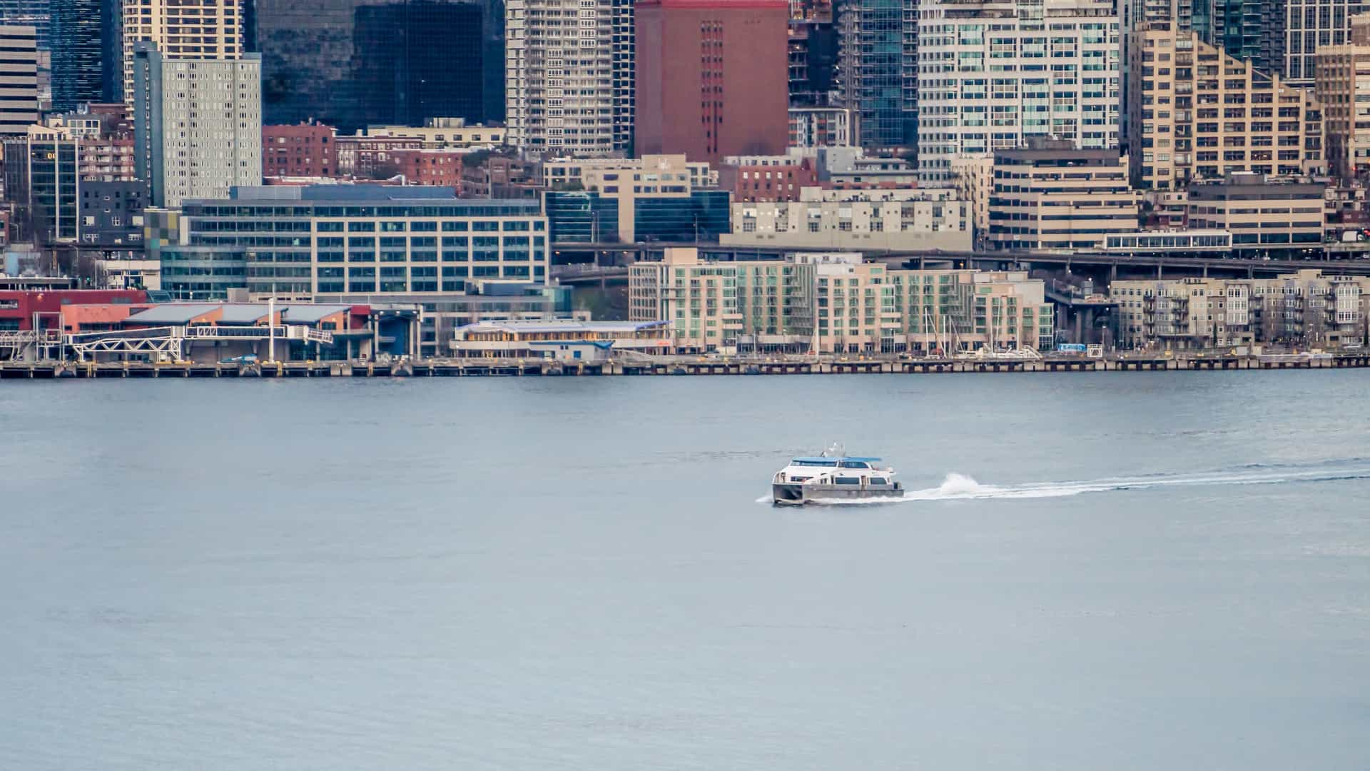 A photograph of a boat, likely a ferry or harbor cruise vessel, sailing across the waters of Elliott Bay in Seattle, Washington. The boat is positioned in the foreground, with the dense downtown Seattle skyline visible in the background, which typically includes prominent landmarks such as the Space Needle, the Seattle Great Wheel, and numerous skyscrapers.