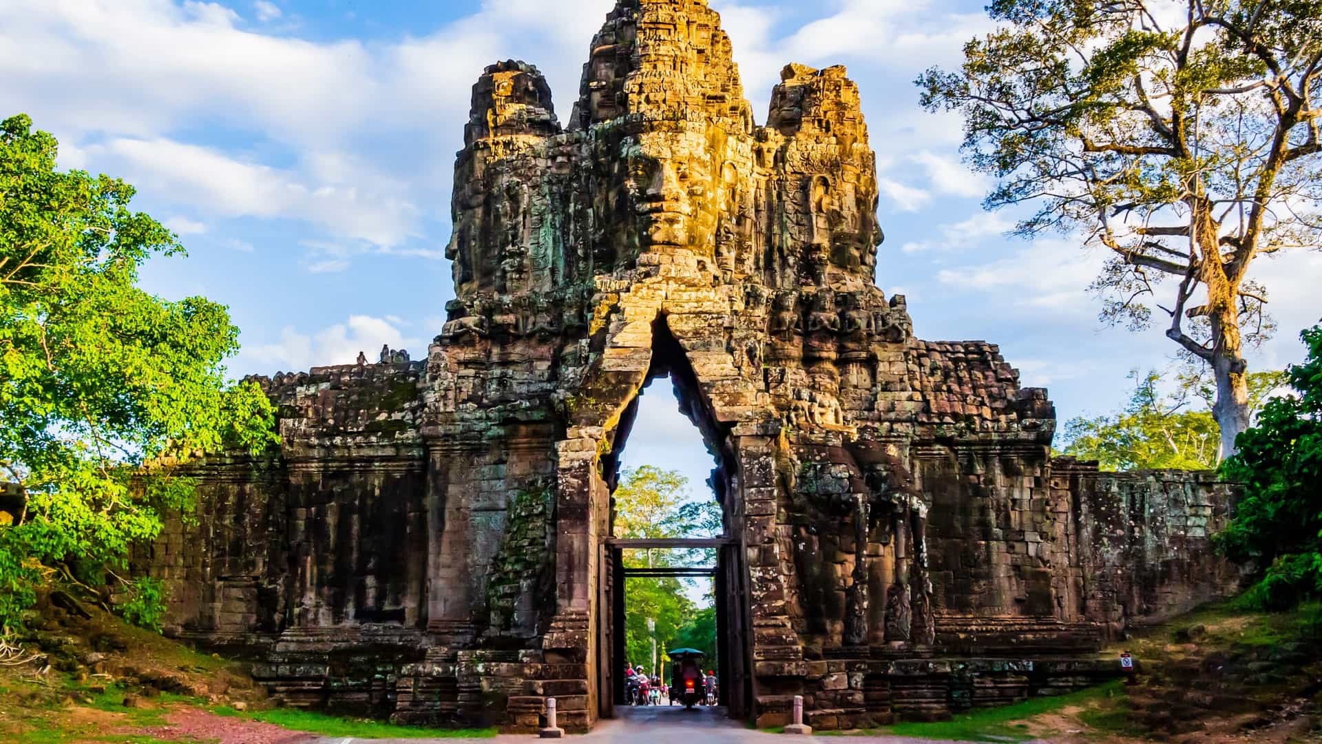 A massive, ancient stone gateway, likely one of the entrances to Angkor Thom, stands tall with its iconic sculpted faces looking out. The dramatic structure, built of dark, weathered stone, is partially framed by bright green foliage. The gate opens onto a path where a few small vehicles or people are visible, all located near Siem Reap, Cambodia.