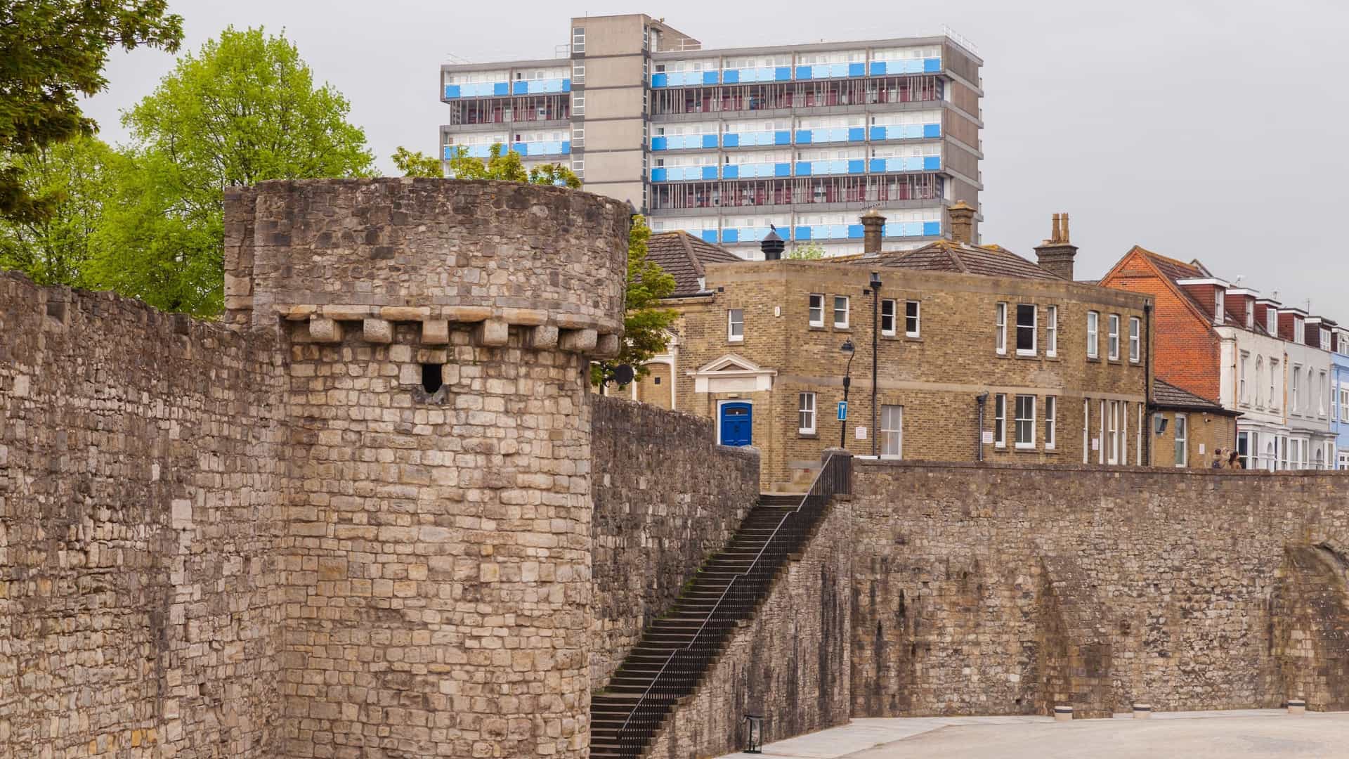  A view along a surviving section of the medieval Southampton Old Town Walls, showing the stone defensive structure with a rounded flanking tower, possibly the Arundel or Catchcold Tower. The historic stone wall contrasts with the modern grass and pavement in the foreground.