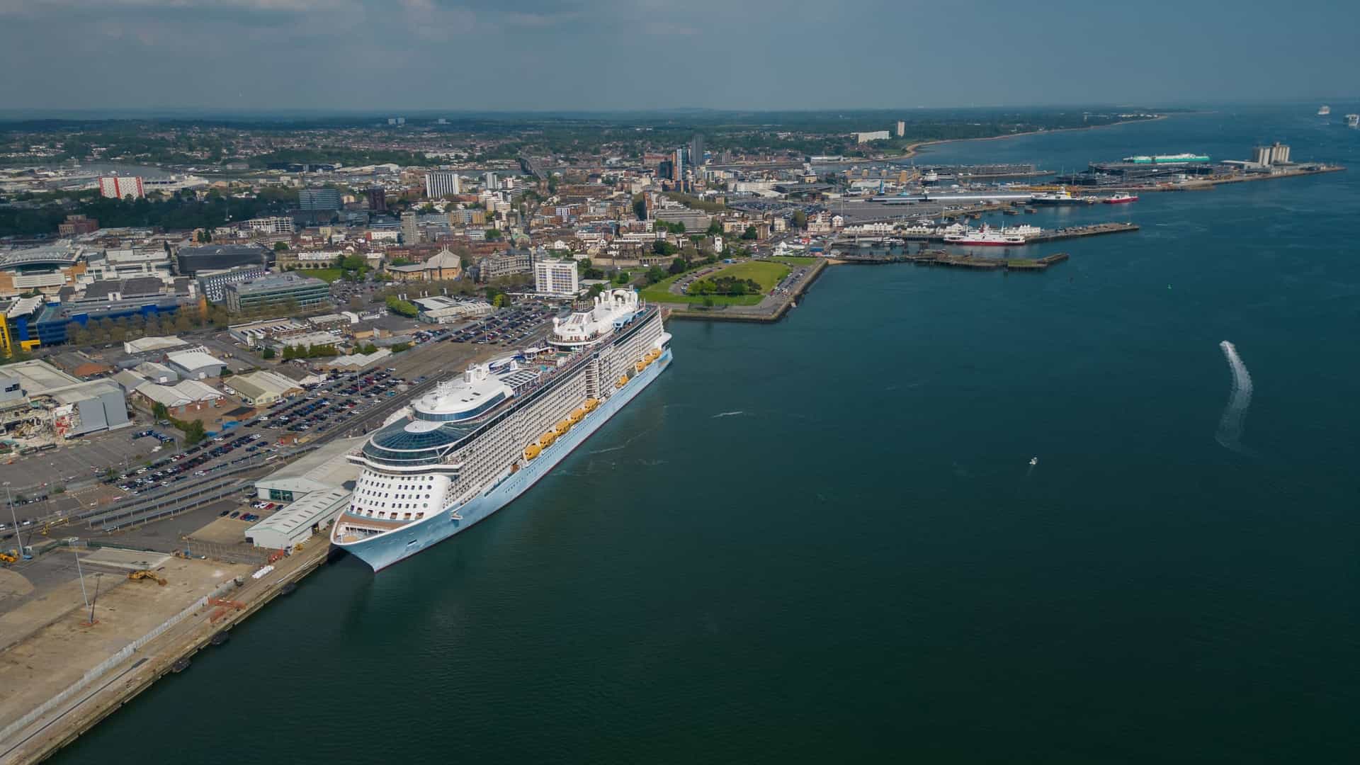  A photograph of a massive, multi deck cruise ship, likely a modern vessel, moored at a dock in the port of Southampton, UK. The image captures the scale of the ship against the port infrastructure and the surrounding sky or city backdrop.