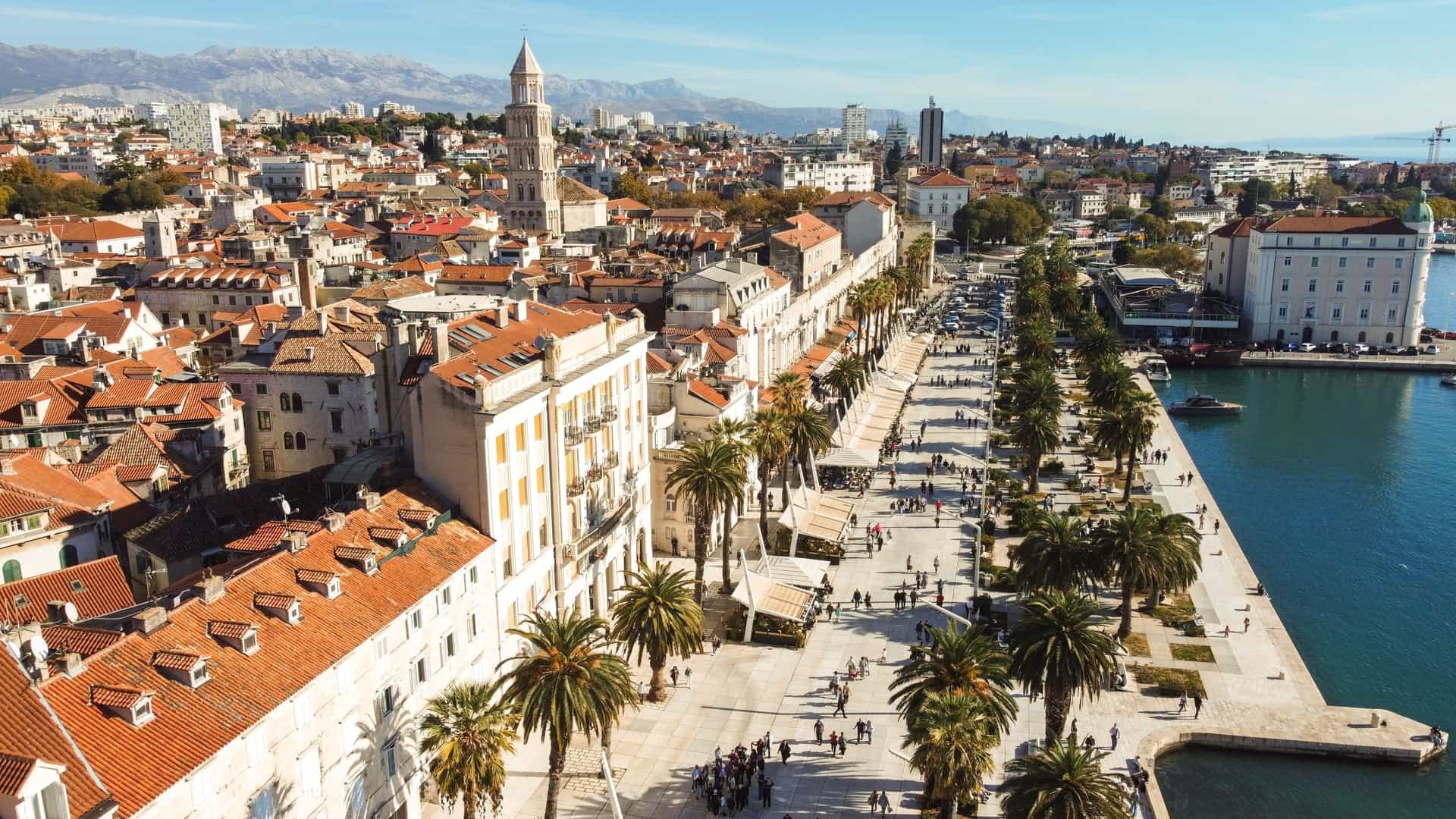  A high angle aerial view of the Riva waterfront promenade in Split, Croatia, bordering the Adriatic Sea. The image prominently features the historic stone walls of Diocletian's Palace (a UNESCO World Heritage site) and its red tiled rooftops on the right, contrasted with the palm tree lined promenade and the bustling harbor with boats on the left. The cityscape is dense, with the blue water of the sea completing the scene.