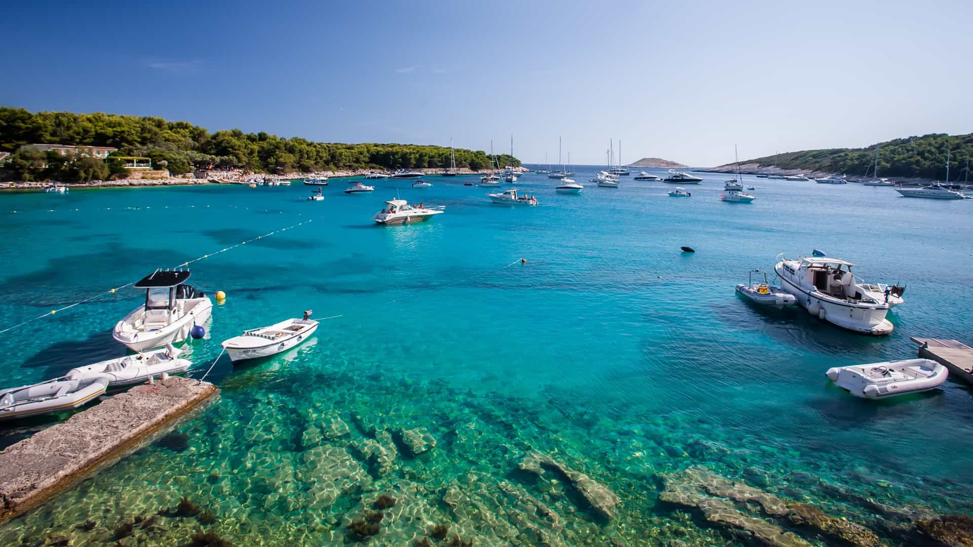 A scenic view of the harbor in Hvar Town, on the island of Hvar, Croatia. The image captures several yachts, sailing boats, and smaller vessels moored along a stone waterfront. The background features the historic, red roofed buildings of the town and may include the hills and the prominent Fortica fortress. The water of the Adriatic Sea is typically clear and blue.