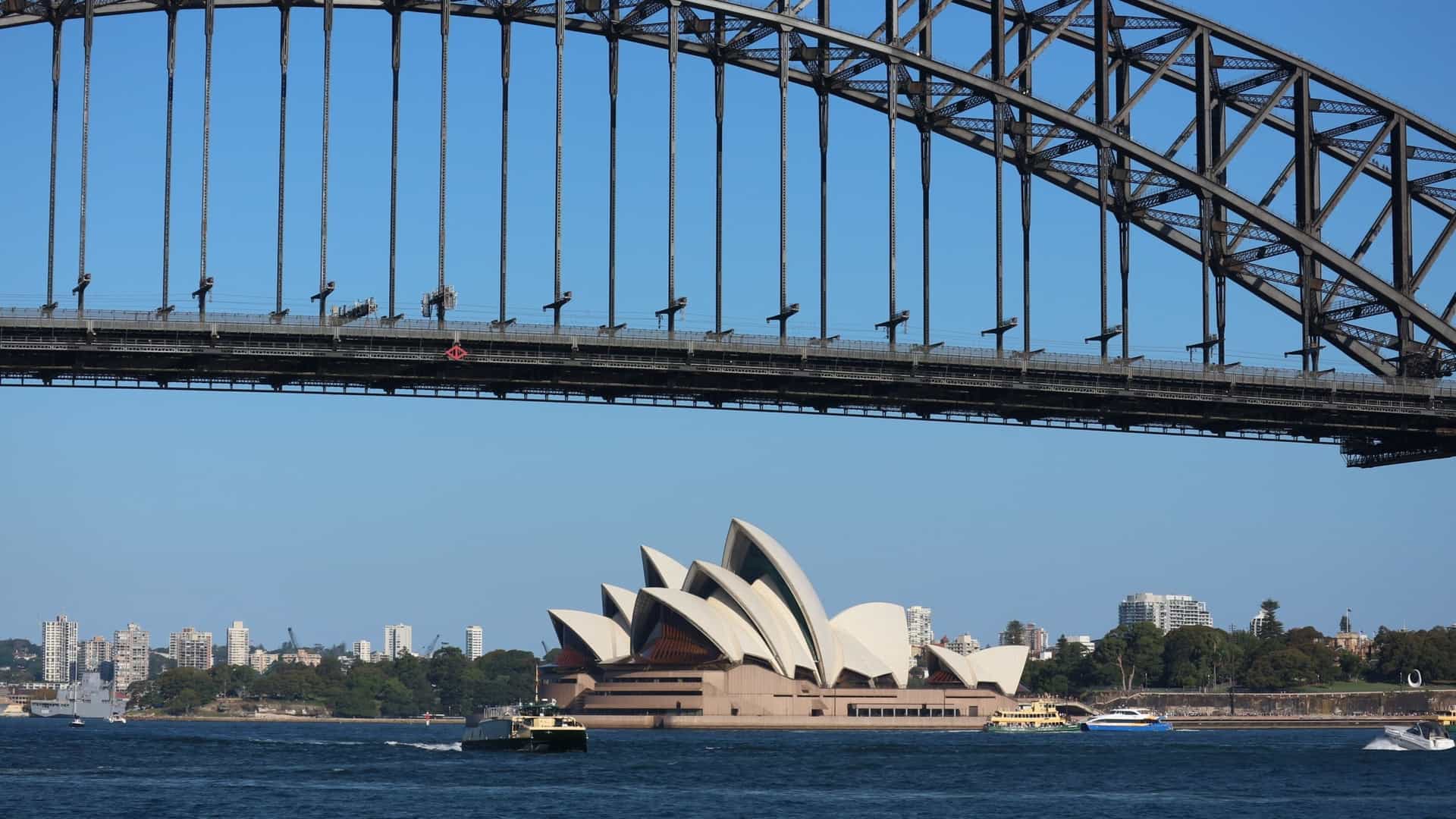 A panoramic view of Sydney Harbour showcasing the iconic Sydney Opera House with its white, sail shaped shells in the foreground, and the steel arch of the Sydney Harbour Bridge spanning the water behind it. The scene is likely captured at sunset or dusk, with reflections on the water and a vibrant city skyline in the background.