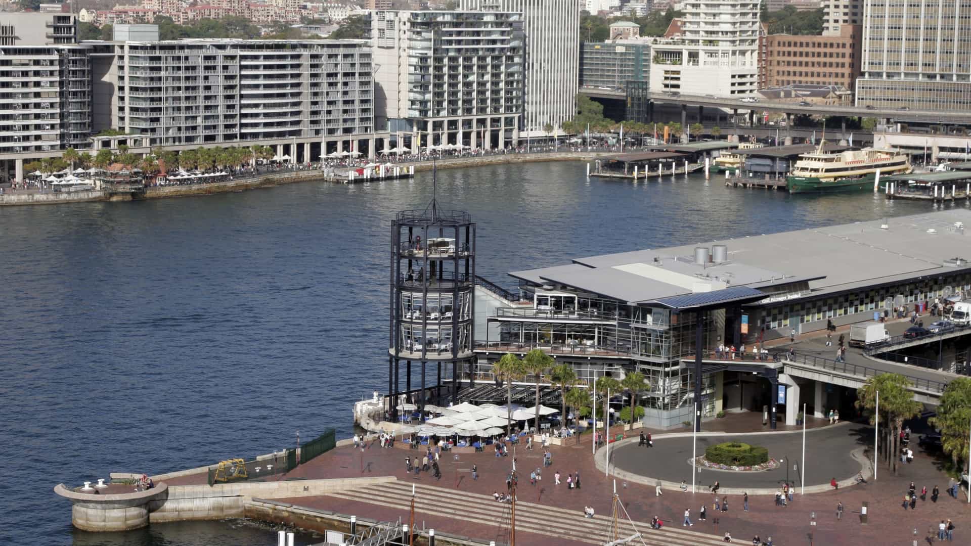  A view of Circular Quay West in Sydney, Australia, with historic and modern buildings lining the waterfront. In the foreground, the colonial era sandstone buildings of The Rocks district, including what is now the Museum of Contemporary Art (MCA), are visible. In the background, modern glass and steel high rise office towers of the Sydney Central Business District (CBD) rise, highlighting the juxtaposition of old and new architecture near the harbor.