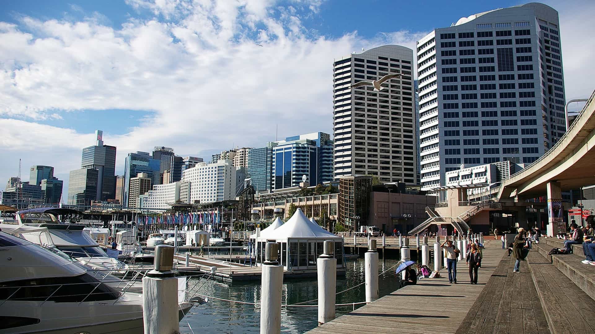  A modern cityscape of Darling Harbour, Sydney, Australia, showing the waterfront precinct. The image features high rise commercial and residential buildings, including the Sydney CBD skyline, surrounding the harbor water, with pleasure boats docked in the bay.