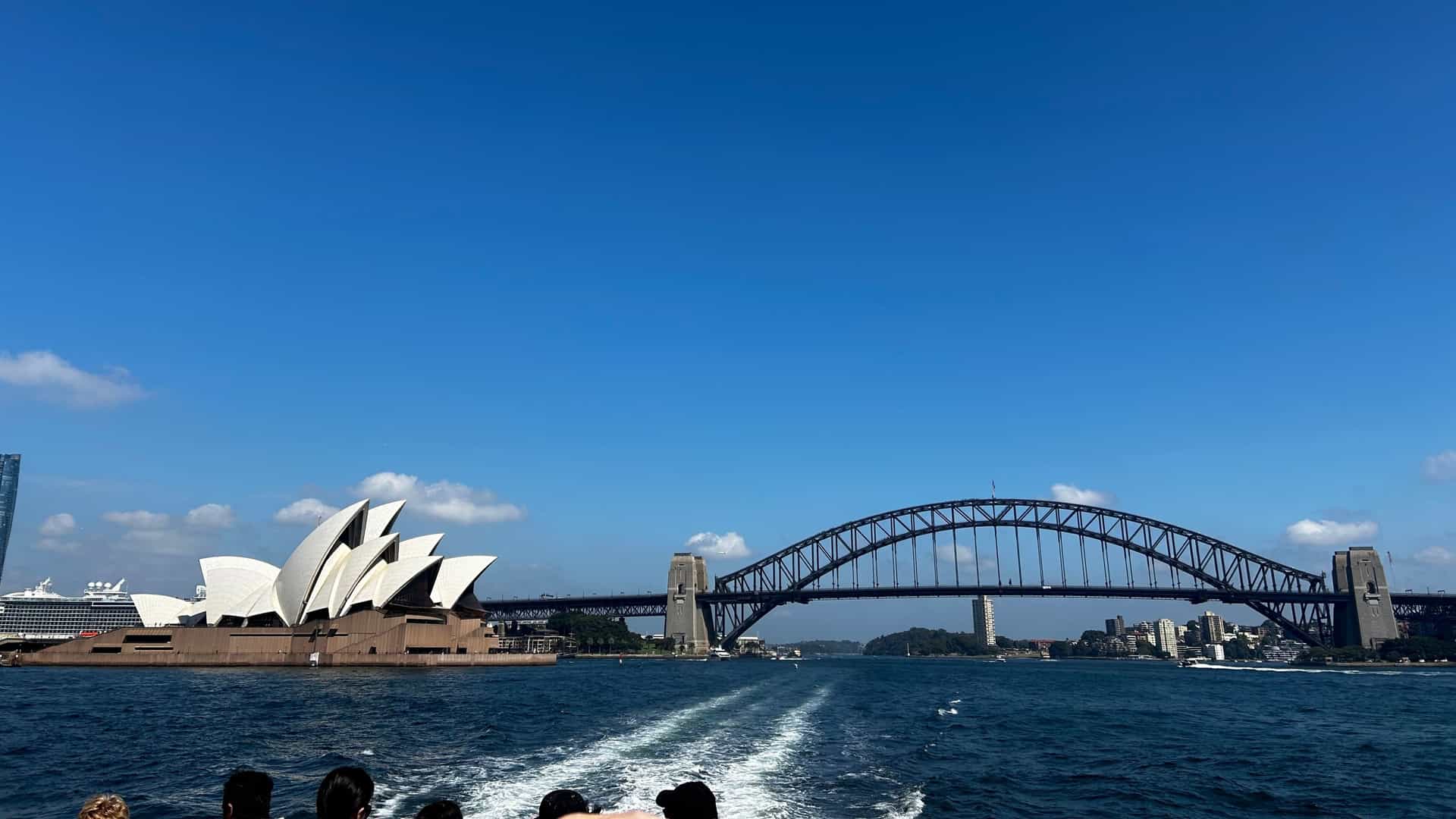  A scenic view of Sydney Harbour with a large cruise ship sailing through the water. In the background, the iconic Sydney Harbour Bridge is visible, along with the dense skyline of the Sydney central business district (CBD) under a bright, clear sky.