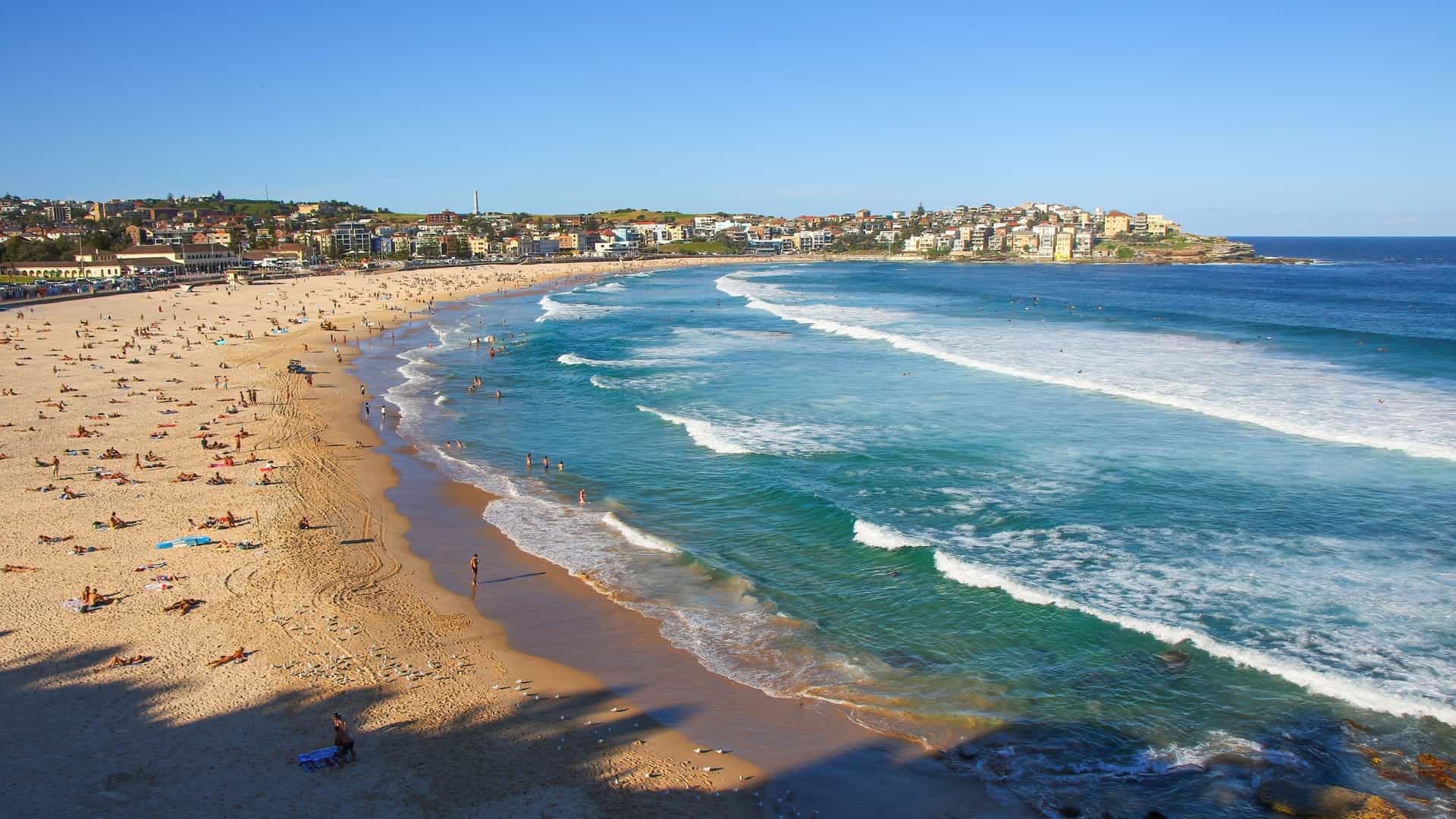  An aerial, high angle view of Bondi Beach in Sydney, New South Wales, Australia, showcasing the long crescent of golden sand, the turquoise blue Pacific Ocean with breaking waves, and the surrounding urban area. The image captures the famous beach and coastline under a bright, sunny sky.