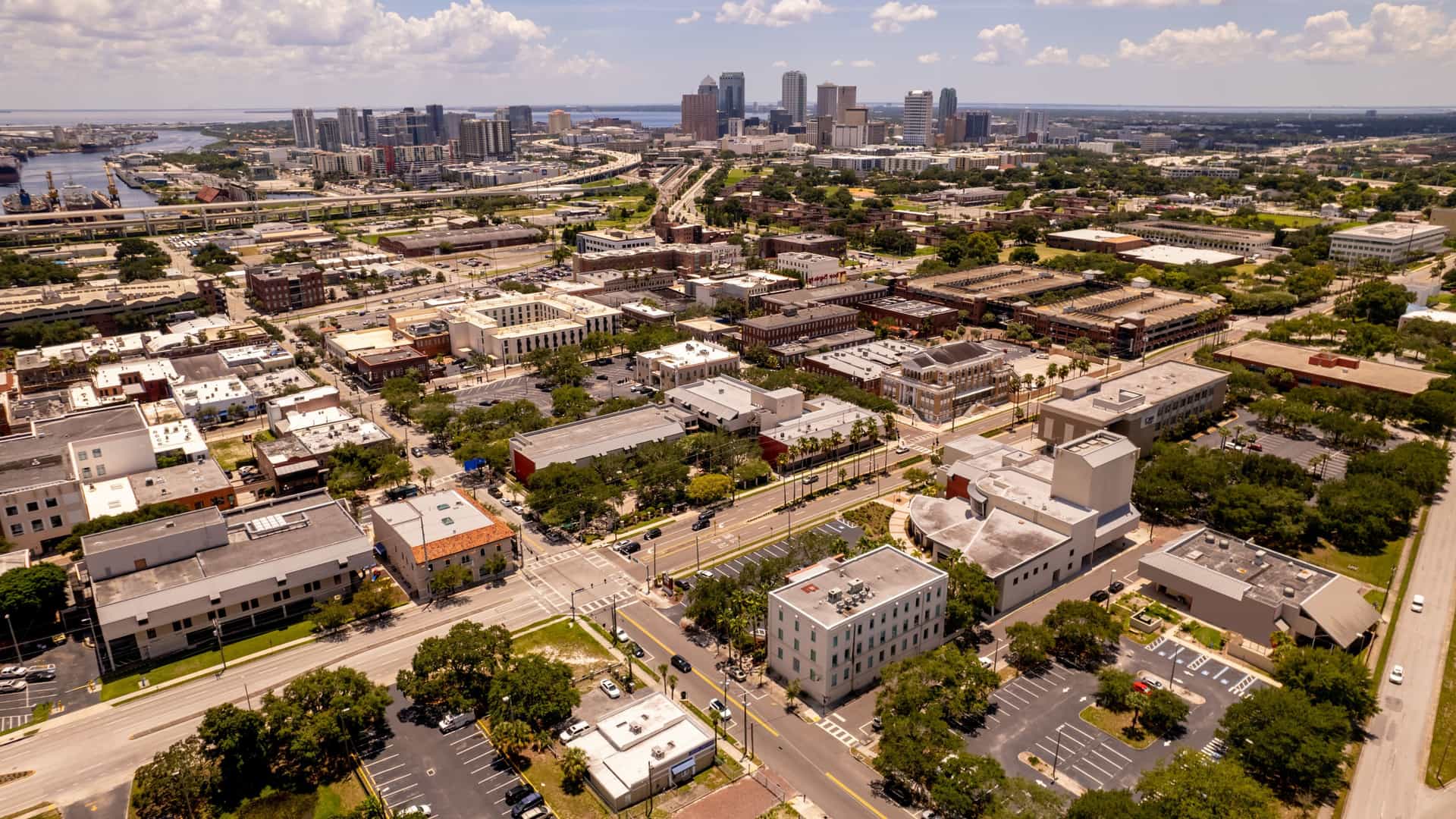 An aerial, panoramic view of Ybor City, the historic Latin Quarter of Tampa, Florida, showing the low-rise, historic architecture in the foreground, with the modern, high-rise skyscrapers of the downtown Tampa skyline visible in the distance.