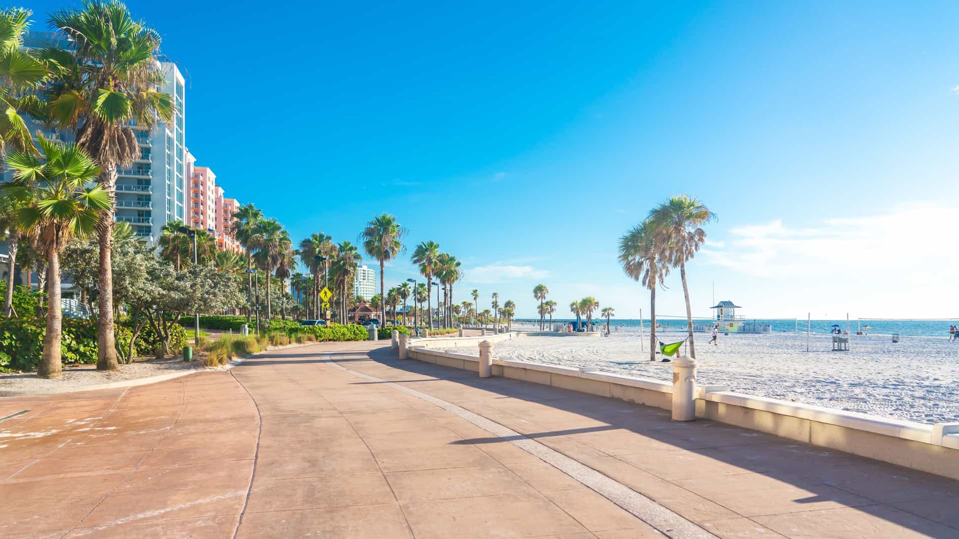 A wide, sunny view of Clearwater Beach in Florida, showcasing its famous, sugar-white quartz sand and the clear, emerald-green water of the Gulf of Mexico. The beach is lined with colorful rental chairs and umbrellas, with a few palm trees and modern beachfront hotels visible in the background.