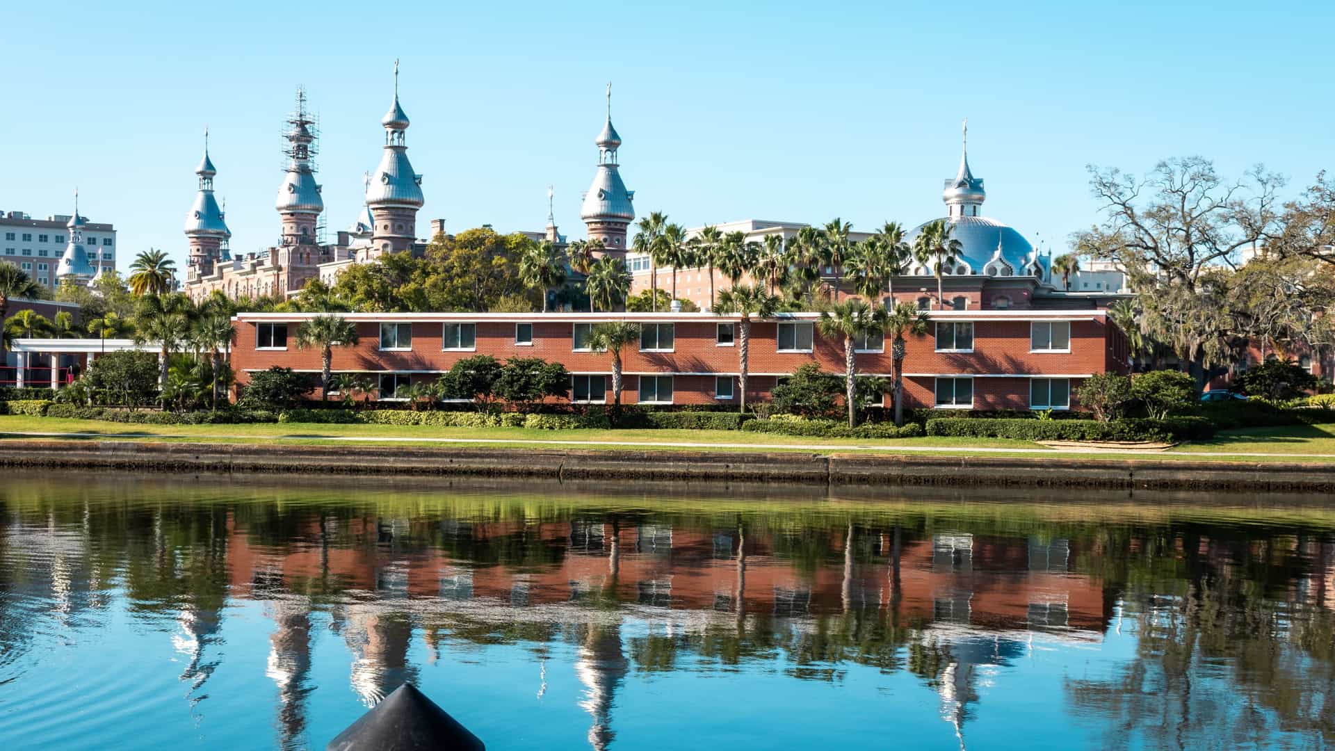 The Henry B. Plant Museum, with its ornate Moorish-Byzantine architecture featuring distinctive minarets and domes, is perfectly reflected in the still waters of the Hillsborough River in Tampa, Florida. Green trees and landscaping line the riverbank in the foreground.