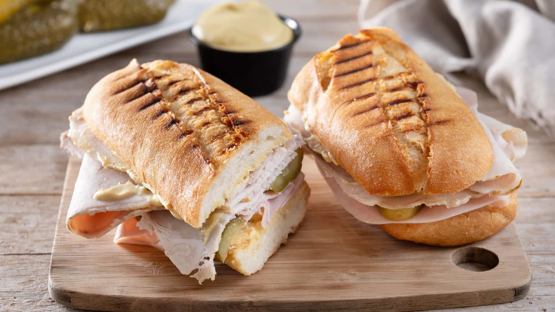A grilled and pressed traditional Cuban sandwich, or Cubano, is cut diagonally and placed on a rustic wooden table. The sandwich consists of Cuban bread, sliced roast pork, ham, Swiss cheese, dill pickles, and yellow mustard, with the crispy, golden-brown bread showing the press marks.