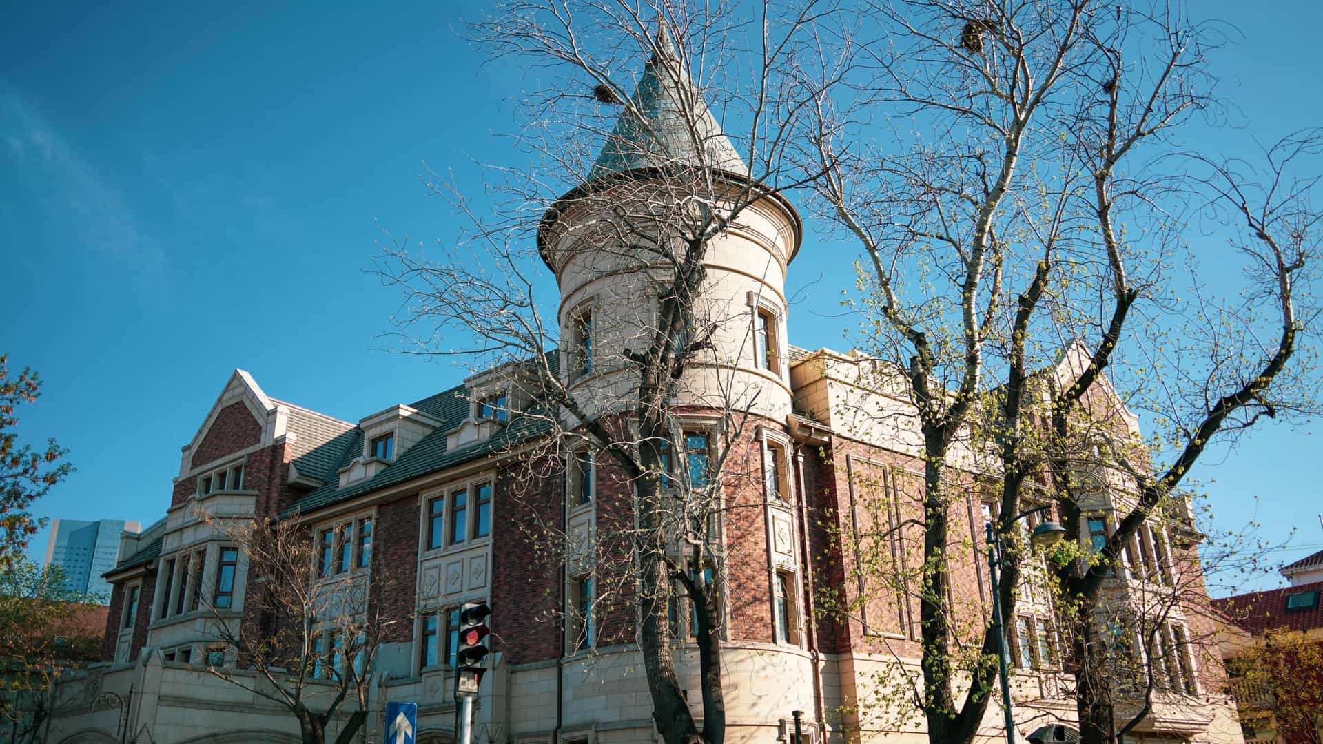  A scenic view of the "Five Great Avenues" (Wudadao) in Tianjin, China, showcasing European style colonial architecture along a tree lined street. The building in the foreground is a historic brick mansion with a red roof, surrounded by green foliage, under a bright sky. The street is wide and quiet, highlighting the district's blend of history and foreign architectural styles.