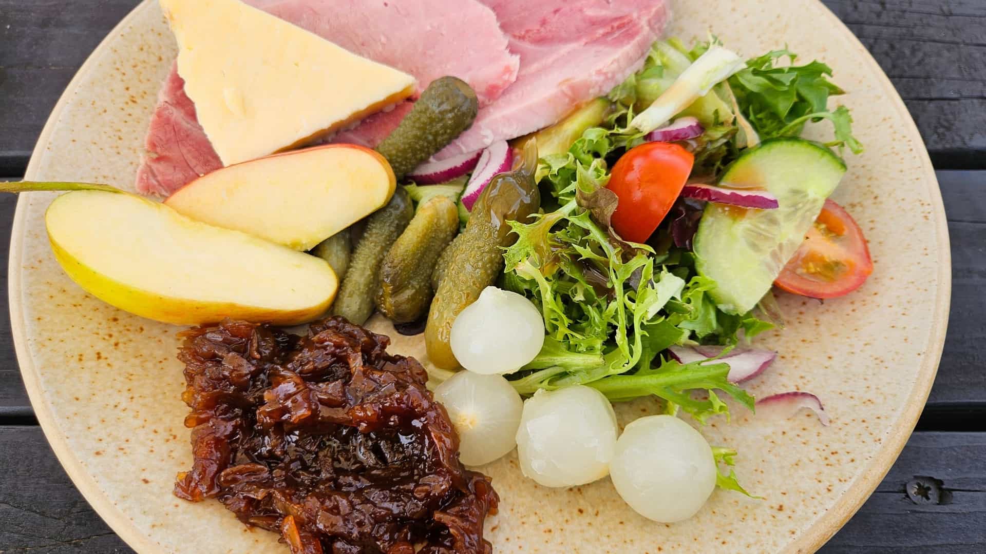  A photograph of a traditional Ploughman's Lunch, a cold British pub meal. The plate or wooden platter typically features a main arrangement of items such as chunks of cheese (like cheddar), a crusty bread roll, and pickled onions or chutney. It may also include other accompaniments like a slice of ham or pork pie, an apple slice, a hard boiled egg, and a simple side salad of lettuce and tomato. The scene is often rustic, befitting its traditional setting in a place like Polstead, Suffolk, England.