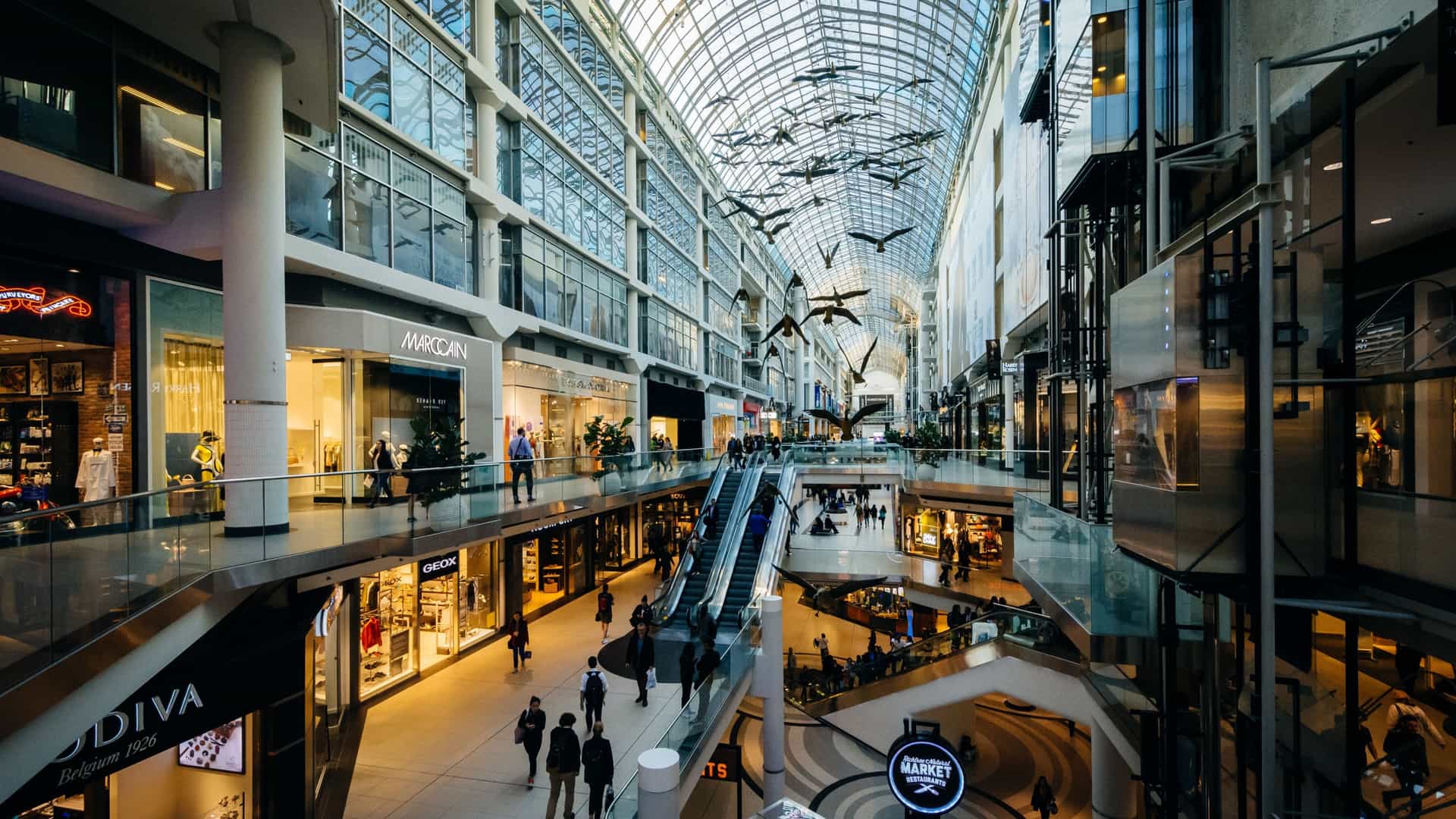  The interior of the multi level CF Toronto Eaton Centre shopping mall in downtown Toronto, Ontario, is shown, highlighting its modern glass galleria architecture and long skylight roof. Shoppers are visible on the different levels, and the famous "Flight Stop" sculpture of fiberglass Canadian geese may be seen hanging from the ceiling.