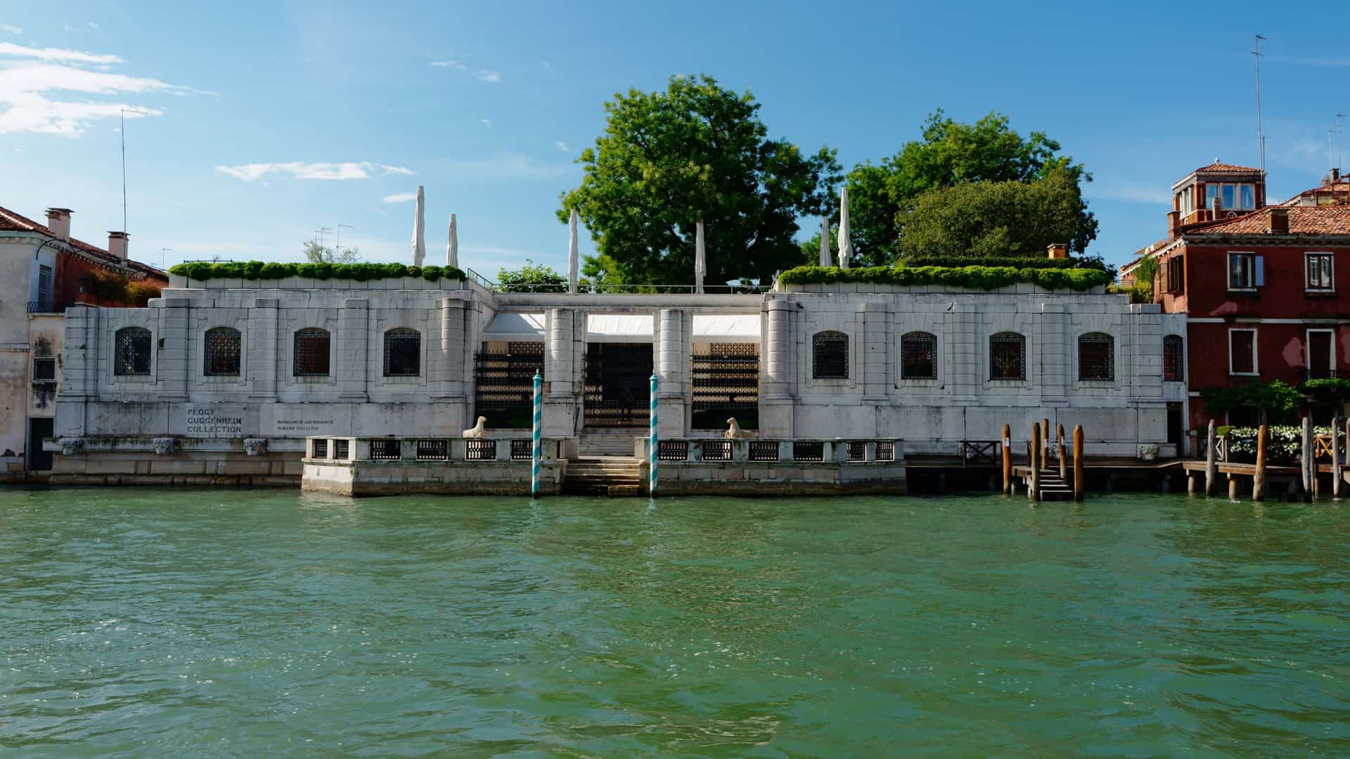  The low, white Istrian stone facade of the Palazzo Venier dei Leoni, home to the Peggy Guggenheim Collection, is seen from the Grand Canal in Venice. The single story palace features an open terrace overlooking the water, a distinctive feature that sets it apart from the taller, multi story palazzi flanking it. The building is situated behind a stone barrier, and boats are visible passing on the canal.
