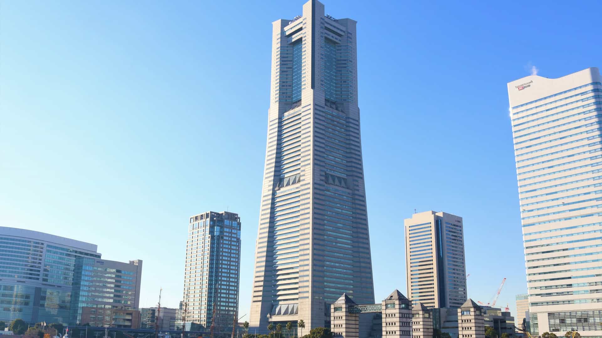 A photograph capturing the Yokohama Landmark Tower and the surrounding Minato Mirai 21 district in Yokohama, Japan. The image likely features the towering skyscraper's distinct architecture against the sky, with other modern buildings, possibly including the Queen's Square Yokohama and the Cosmo Clock 21 Ferris wheel, visible in the vibrant, waterfront urban landscape. The scene may be captured during the day or at night with city lights.