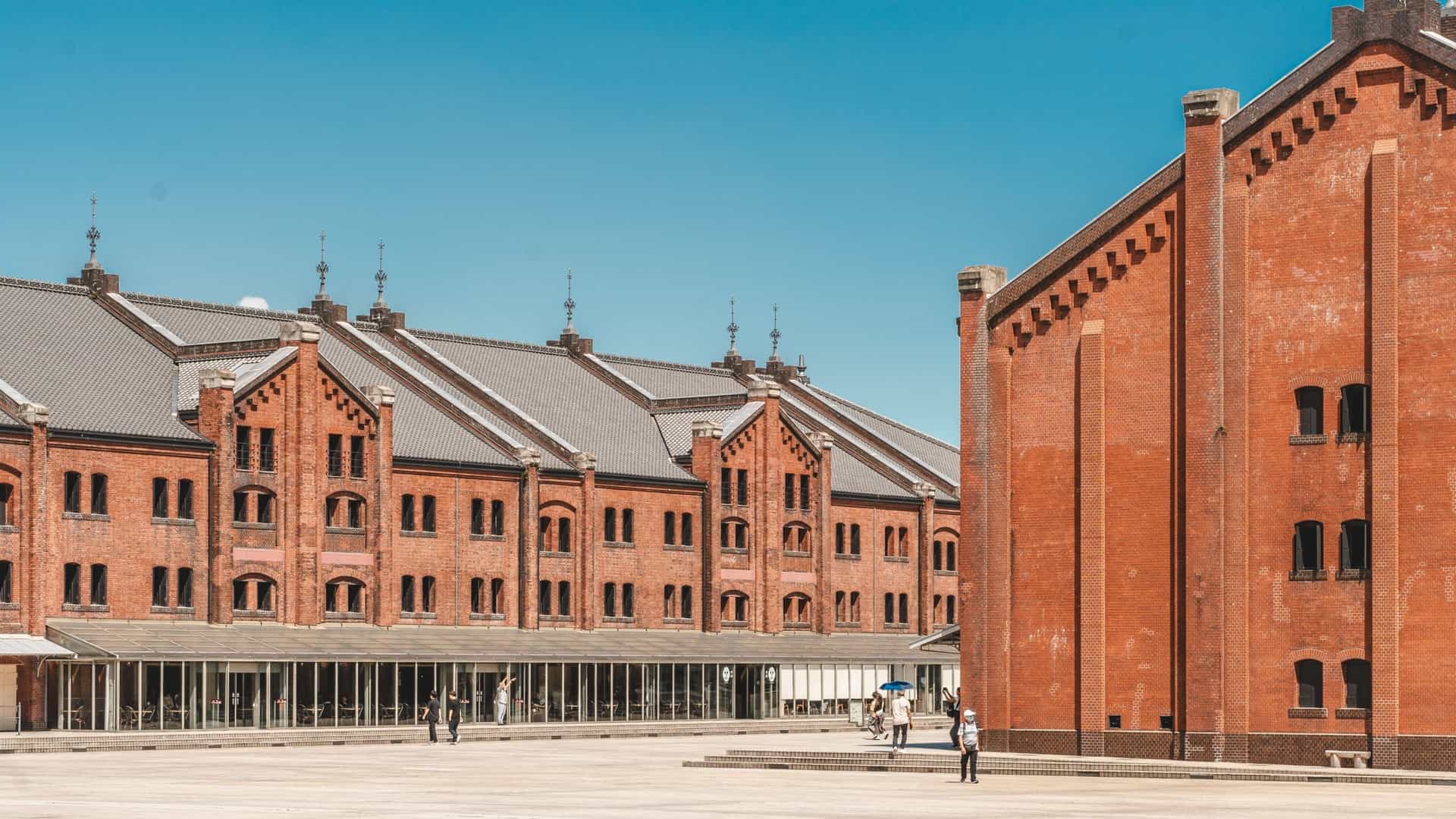  A daytime view of the two historical Yokohama Red Brick Warehouse (Akarenga Sōko) buildings, featuring their distinctive red brick facades and long, rectangular shapes, separated by a paved plaza. The buildings are located along the waterfront, with the modern skyline of the Yokohama Minato Mirai district visible in the background.