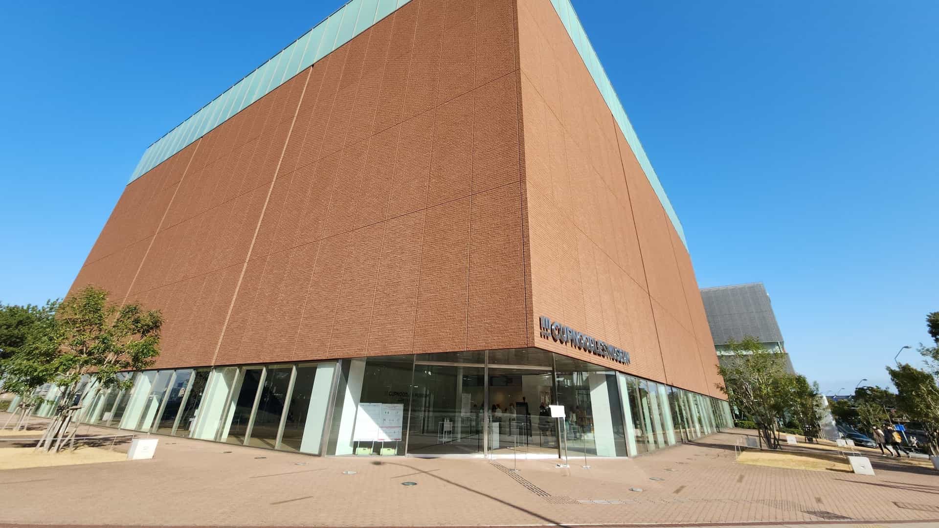  A bright, outdoor, eye level photograph of the Cup Noodles Museum in Yokohama, Japan. The building has a modern, minimalist design, primarily featuring a reddish brown brick facade on the lower levels and large, reflective glass windows on the upper levels. The entrance area is visible, and the building's name, possibly in both Japanese and English, is displayed on the exterior. The sky is bright blue, suggesting a clear day.