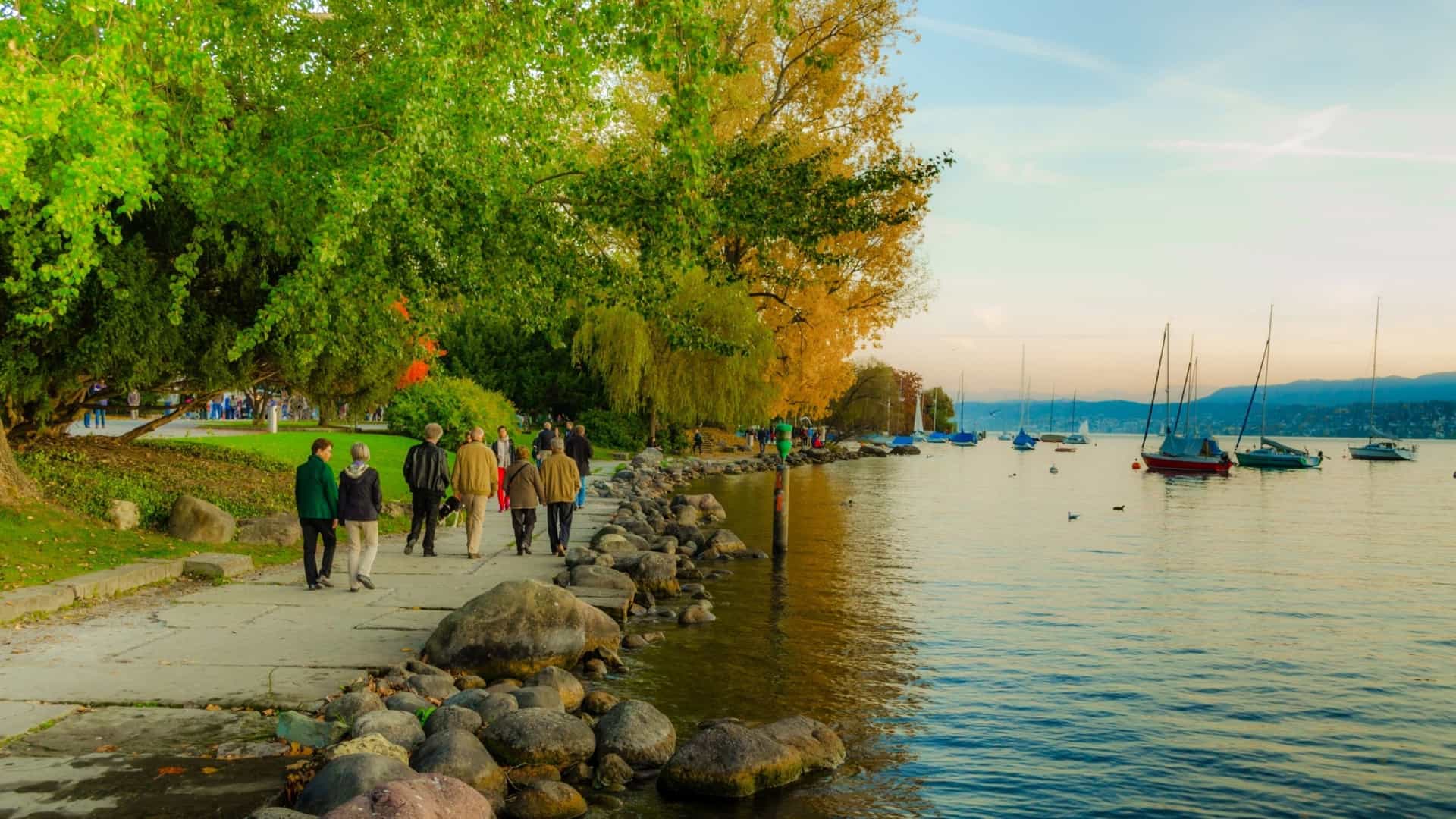  A sunny, slightly blurred, horizontal photograph of a wide coastal promenade with a few people walking. The scene is lined with palm trees and other greenery, suggesting a warm climate, with a view of buildings and the sea in the background.