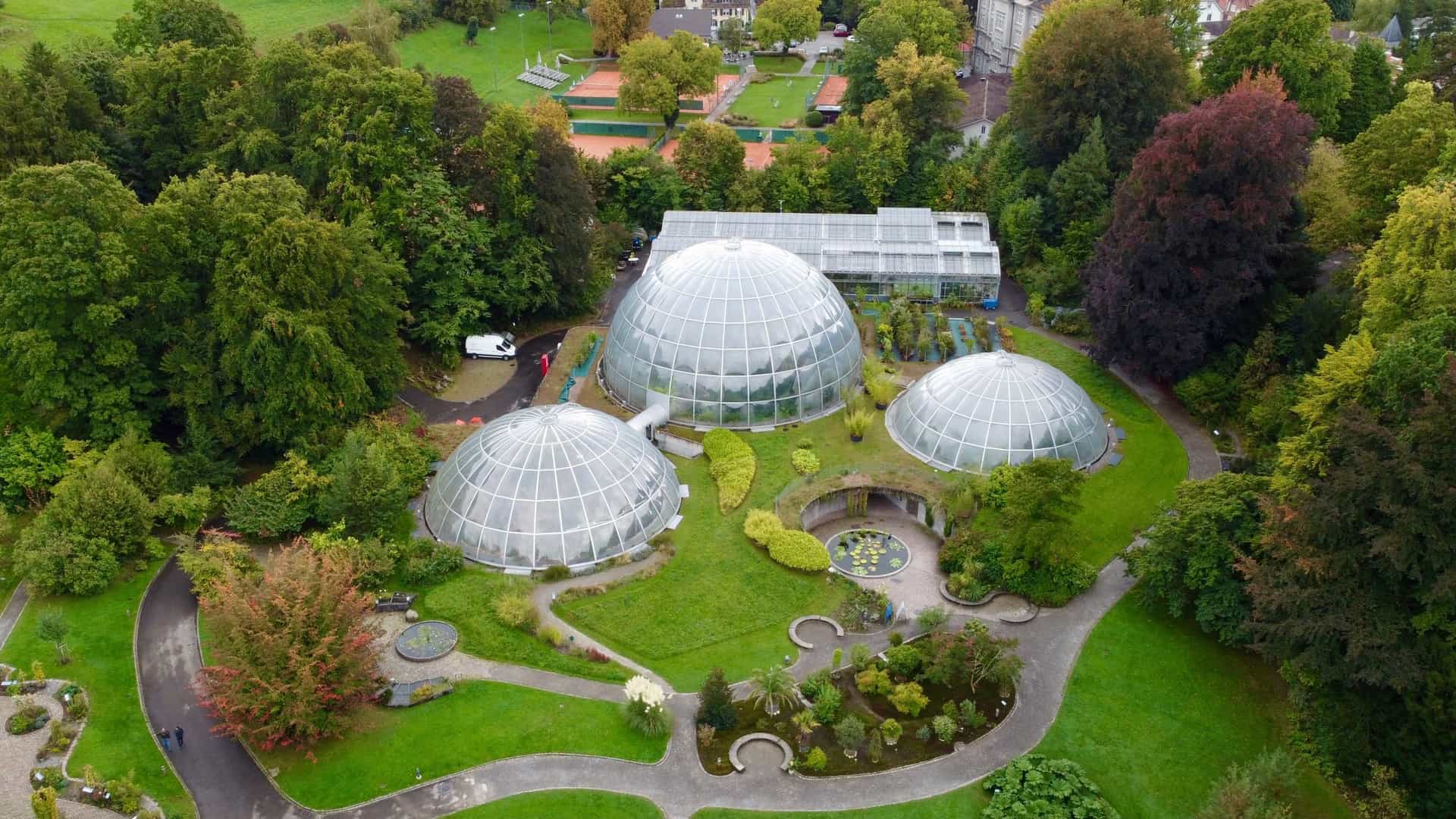  An aerial, top down view of the Botanical Garden of the University of Zurich in Switzerland. The image prominently features the garden's three distinctive, futuristic looking, dome shaped glasshouses (tropical houses) nestled amidst a lush green landscape of trees and various plant collections. The image was captured with a drone (DJI mini2).