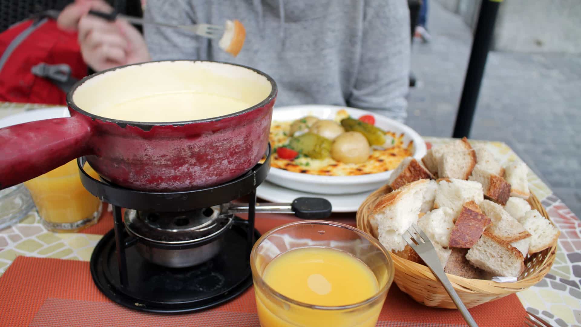  A close up, outdoor photo of a traditional Swiss cheese fondue served at an outdoor café in Lucerne, Switzerland. The image shows the clay fondue pot (caquelon) set atop a small burner, filled with melted cheese, with metal fondue forks dipping pieces of bread into it. The setting, often featuring blankets or a cozy atmosphere, suggests a cold weather meal on a terrace, potentially with a backdrop of Lucerne's historic buildings.