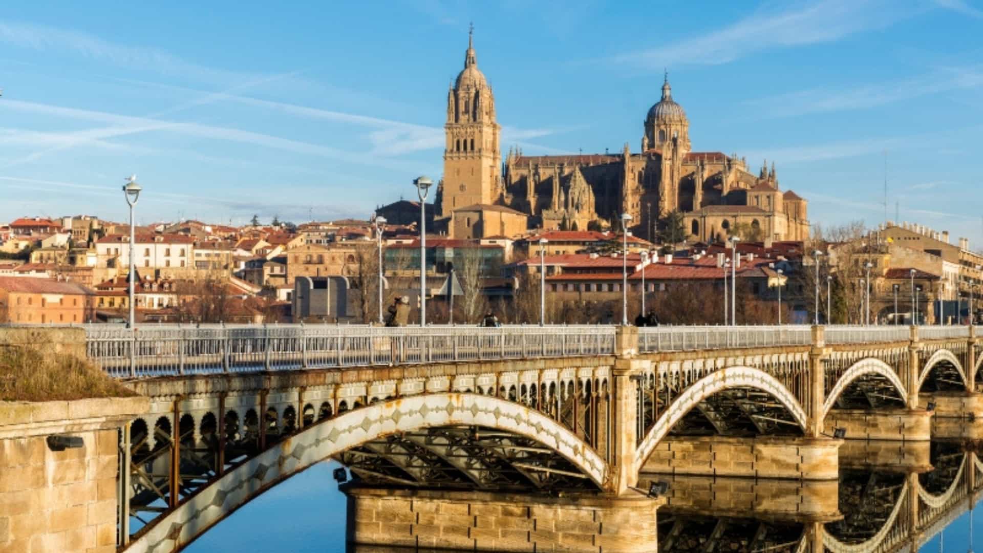 A beautiful view of the city of Salamanca, Spain, located on the Douro River. The foreground is dominated by a historic arched bridge with ornate metalwork, spanning the calm, reflective river. On the opposite bank, the cityscape rises, with the prominent Salamanca Cathedral standing majestically in the background. The cathedral's spires and domes are silhouetted against a bright, clear blue sky with thin clouds. The image captures the historical beauty of a major city on the Douro River.