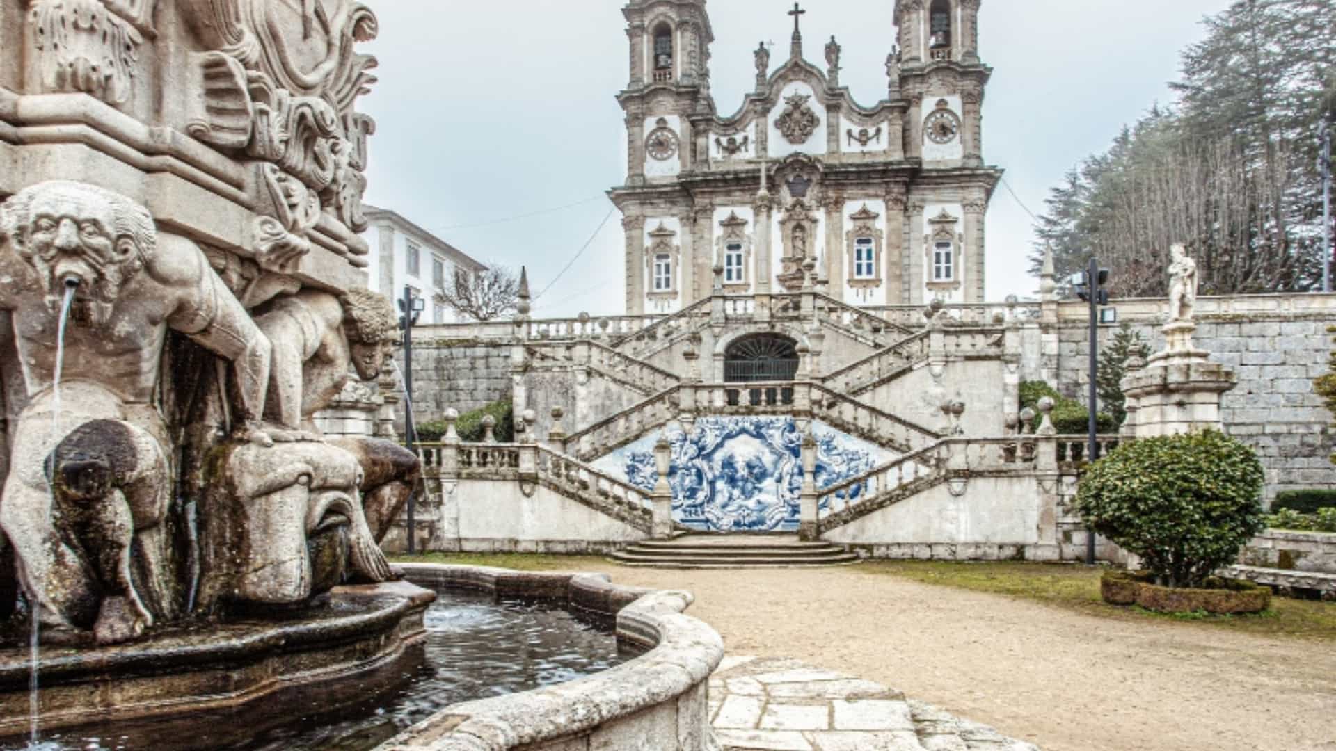 The Sanctuary of Nossa Senhora dos Remédios in Lamego, Portugal, a significant site near the Douro River, is the main focus of this image. A large, ornate stone fountain is in the foreground. In the center, a monumental staircase with blue and white tile panels (azulejos) leads up to the baroque church with its two bell towers. The architecture is grand and intricate, with a stone balustrade lining the steps. The scene is set on a slightly overcast day, which gives the stone and tiles a subtle, muted tone. The image captures a key landmark in the Douro region.
