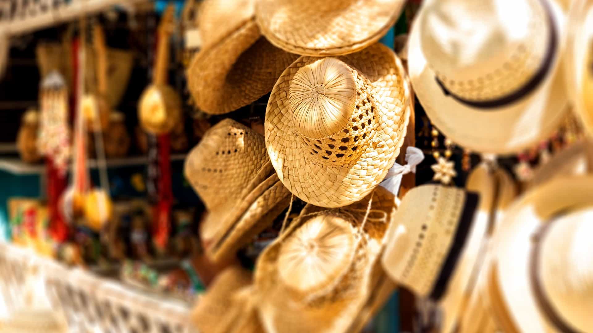 A close-up photograph of a variety of woven straw hats hanging for sale at an outdoor street market in the Eastern Caribbean. The hats are different shapes and sizes, and their intricate textures are clearly visible. In the background, other souvenirs and crafts are blurred, suggesting a lively and colorful market atmosphere. The image captures a quintessential shopping experience on a Caribbean island, a popular activity for visitors.