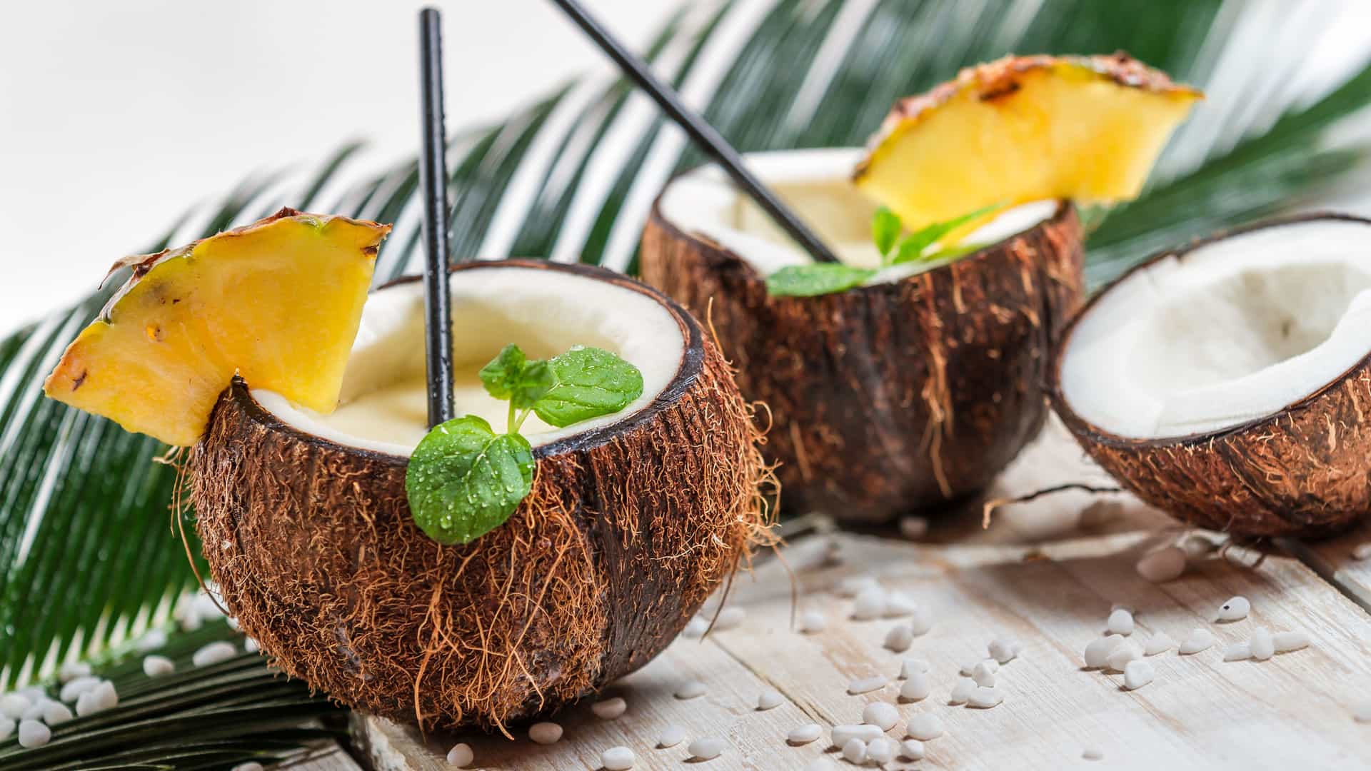 Two delicious piña coladas, a classic tropical drink of the Eastern Caribbean, are served in hollowed-out coconut shells. Each drink is garnished with a fresh pineapple wedge and a sprig of mint. One coconut is cut open on the side, revealing its white interior. The drinks are placed on a light-colored wooden surface scattered with small white pebbles. A large green palm frond is visible in the background, completing the classic tropical theme of this image.