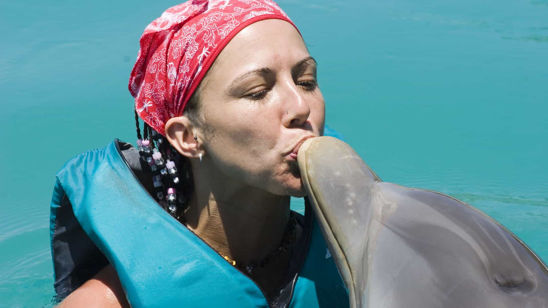 A woman wearing a blue life vest and a red bandana is swimming in the bright turquoise water of the Eastern Caribbean. She is leaning in to kiss the snout of a bottlenose dolphin. The dolphin's skin is a smooth gray color. The woman has a joyous expression on her face, with her eyes closed. This image captures the popular tourist activity of swimming and interacting with dolphins, which is a common experience in the Caribbean region.