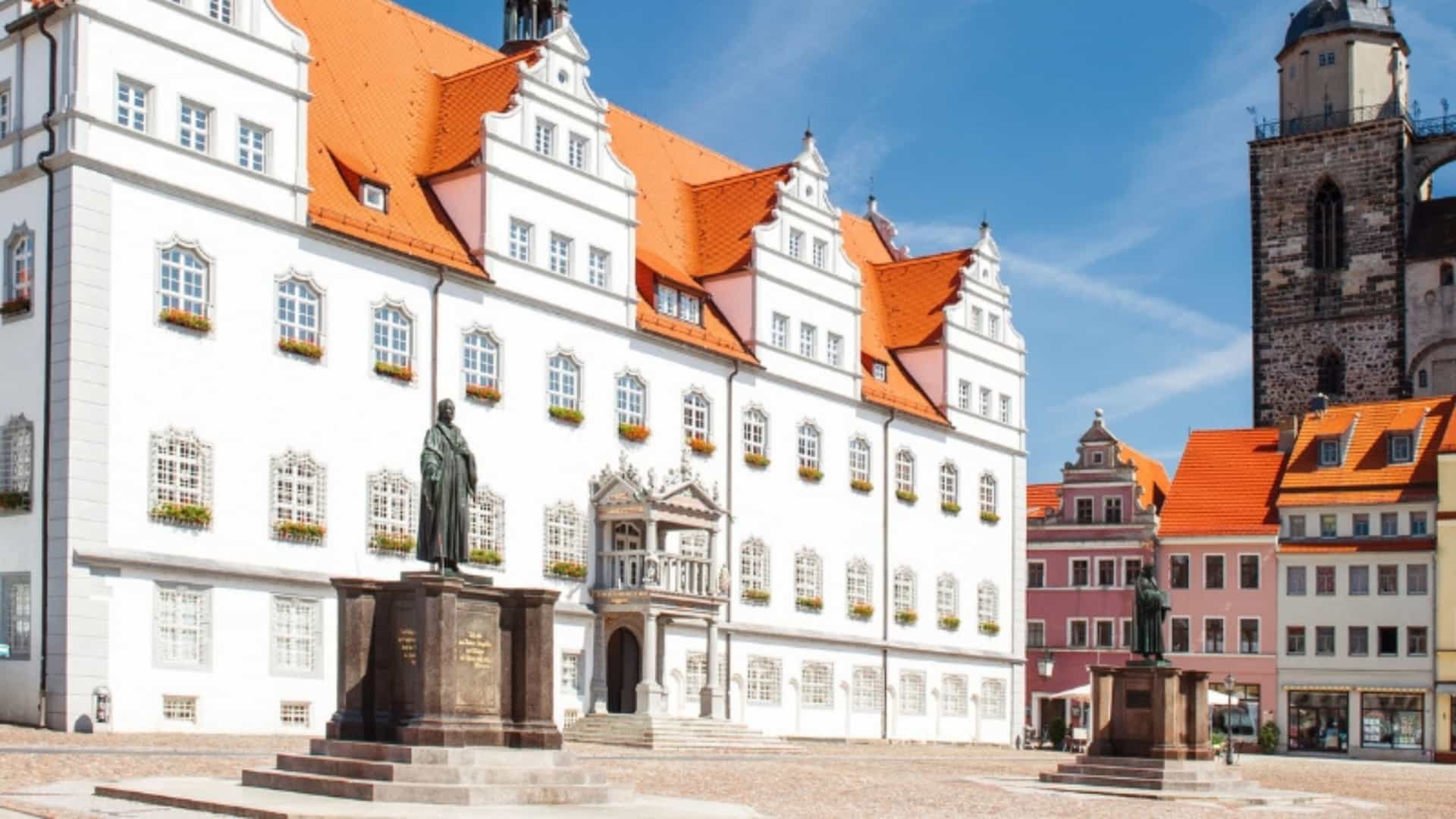 The historic Market Square in Wittenberg, Germany, a city on the Elbe River. The square is dominated by a large, white building with a striking red roof and ornate gables. In front of the building stands a statue of Martin Luther on a stone pedestal. To the right, a second statue is visible, along with the top of a historic church tower. The square is paved with cobblestones, and the sky is a clear, vibrant blue. The image captures the historical and architectural charm of a key city in the Elbe River region.