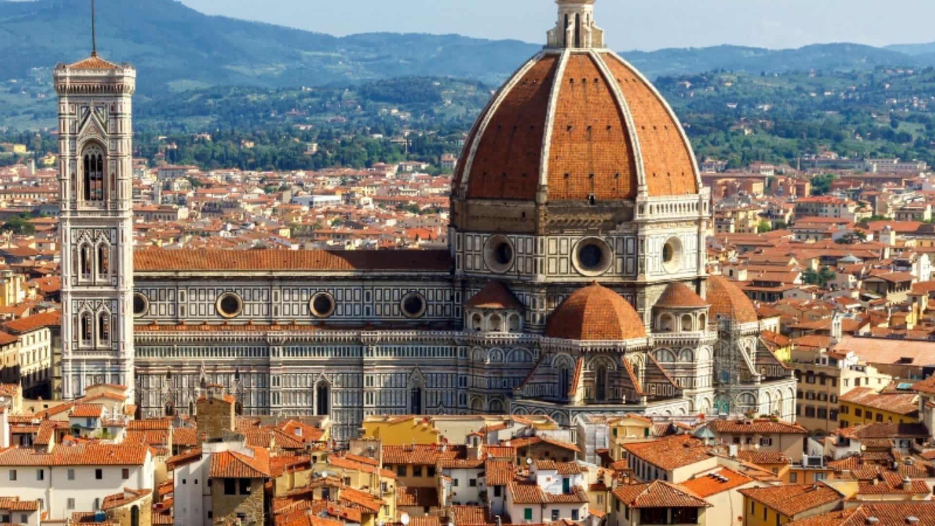 A beautiful aerial view of the Florence Cathedral, or Duomo, and Giotto's Bell Tower in Florence, Italy. The cathedral's iconic red-tiled dome and the intricate patterns of its marble facade are the central focus. The cityscape of Florence, with its sea of red-tiled roofs, stretches out around the landmark. Rolling hills and mountains are visible in the distance under a bright blue sky. This image represents a city often visited as part of a broader European trip, sometimes in conjunction with a river cruise.