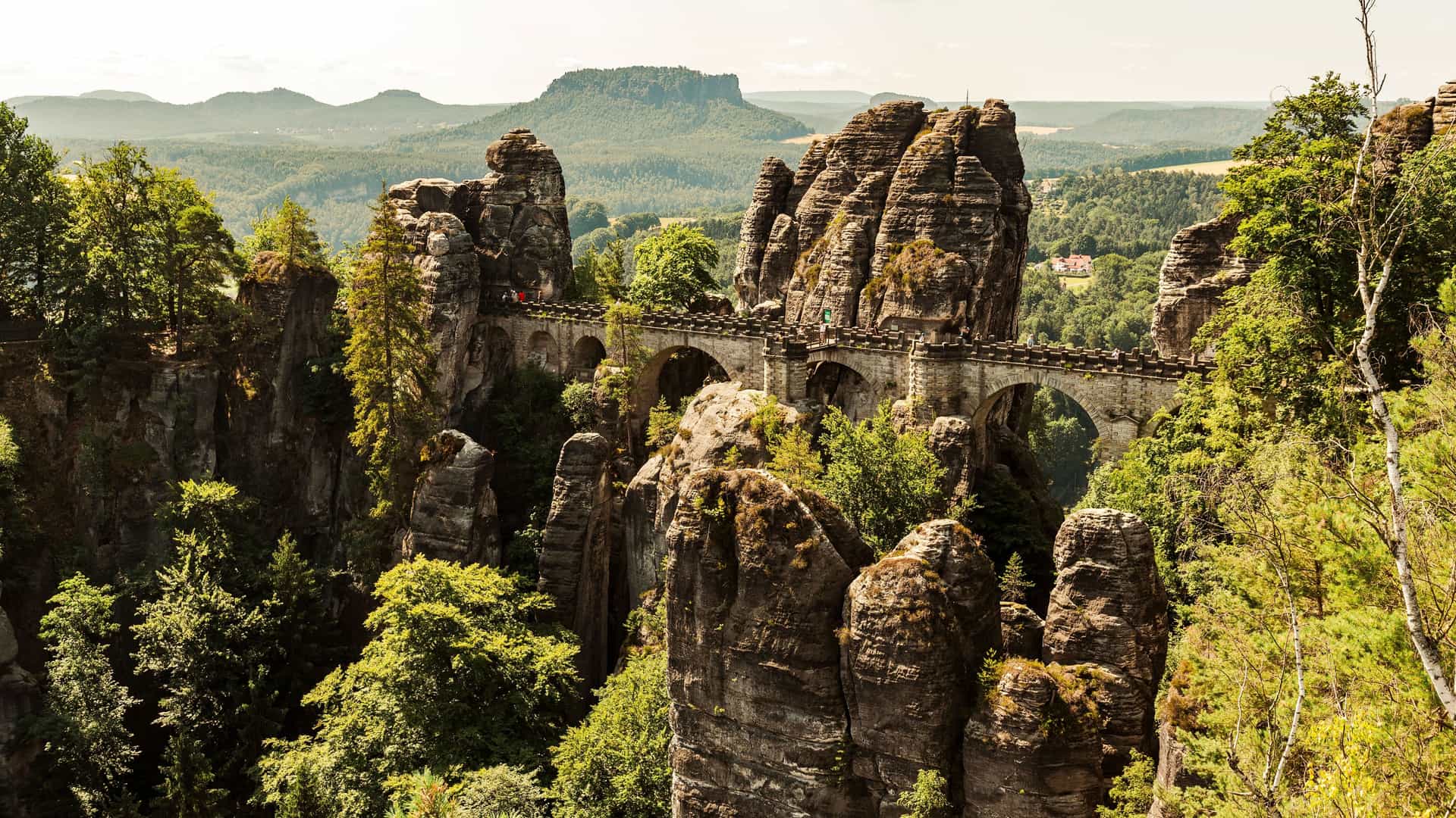 The dramatic Bastei Bridge in Saxon Switzerland, Germany, an area close to the Elbe River. The bridge is made of stone and spans a series of towering, jagged rock formations. Green trees and foliage grow around and on the rocks, adding to the rugged, natural landscape. In the distance, more flat-topped mountains and forests stretch toward the horizon under a cloudy sky. The image captures the impressive and unique natural beauty of the Elbe Sandstone Mountains.