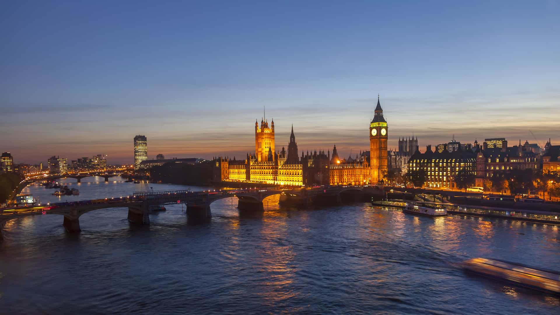 An iconic nighttime view of the London skyline, featuring the illuminated Houses of Parliament and the Elizabeth Tower, commonly known as Big Ben. The golden lights of the buildings reflect on the dark water of the River Thames. The Westminster Bridge, with streetlights glowing, crosses the river in the foreground. In the background, other city lights twinkle under a deep blue evening sky, capturing the famous architecture of the European capital.