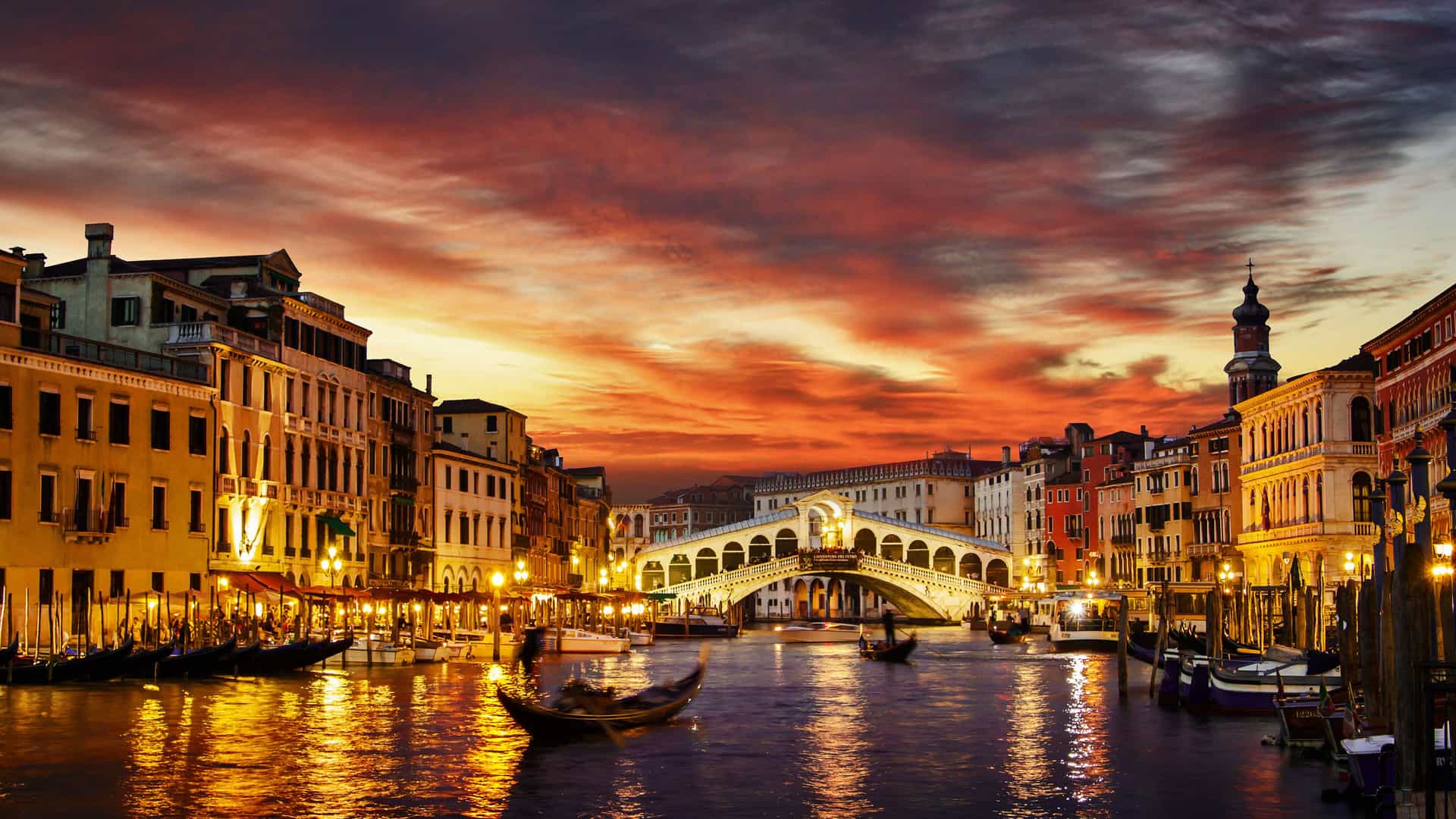 A dramatic sunset over the Grand Canal in Venice, Italy, a famous European destination. The iconic Rialto Bridge stands illuminated in the center, with traditional gondolas and other boats visible on the water. The historic buildings lining the canal are bathed in the warm glow of their lights and the golden-orange light from the sky. The clouds above are a vibrant mix of fiery red and purple, reflecting on the water below, creating a quintessential Venetian scene.